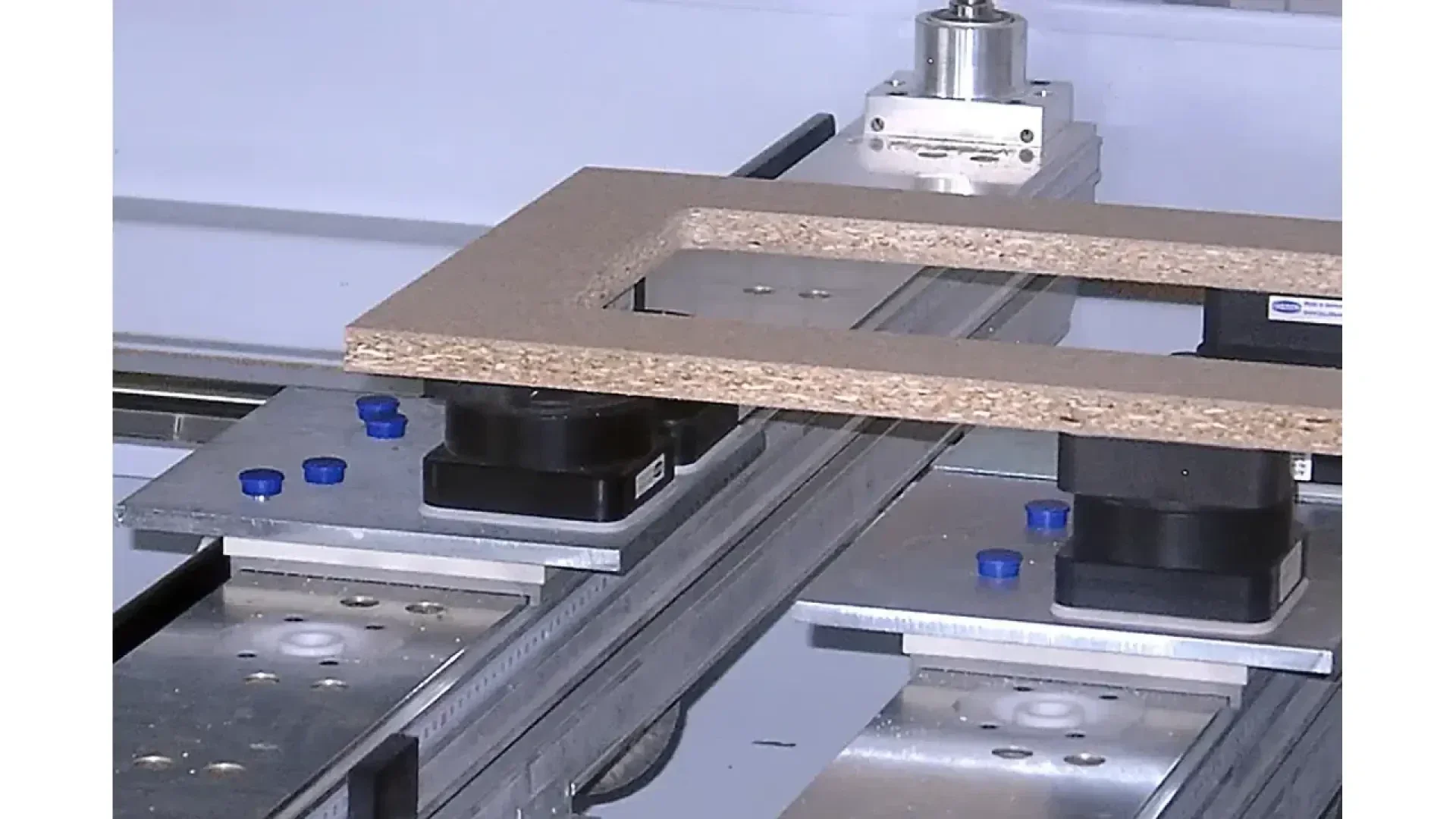 Gros plan d'un cadre rectangulaire en bois placé sur la table d'une machine CNC, avec des rails métalliques, des pinces et des chevilles en plastique bleu visibles, prêt pour les processus de coupe ou de fraisage automatisés.