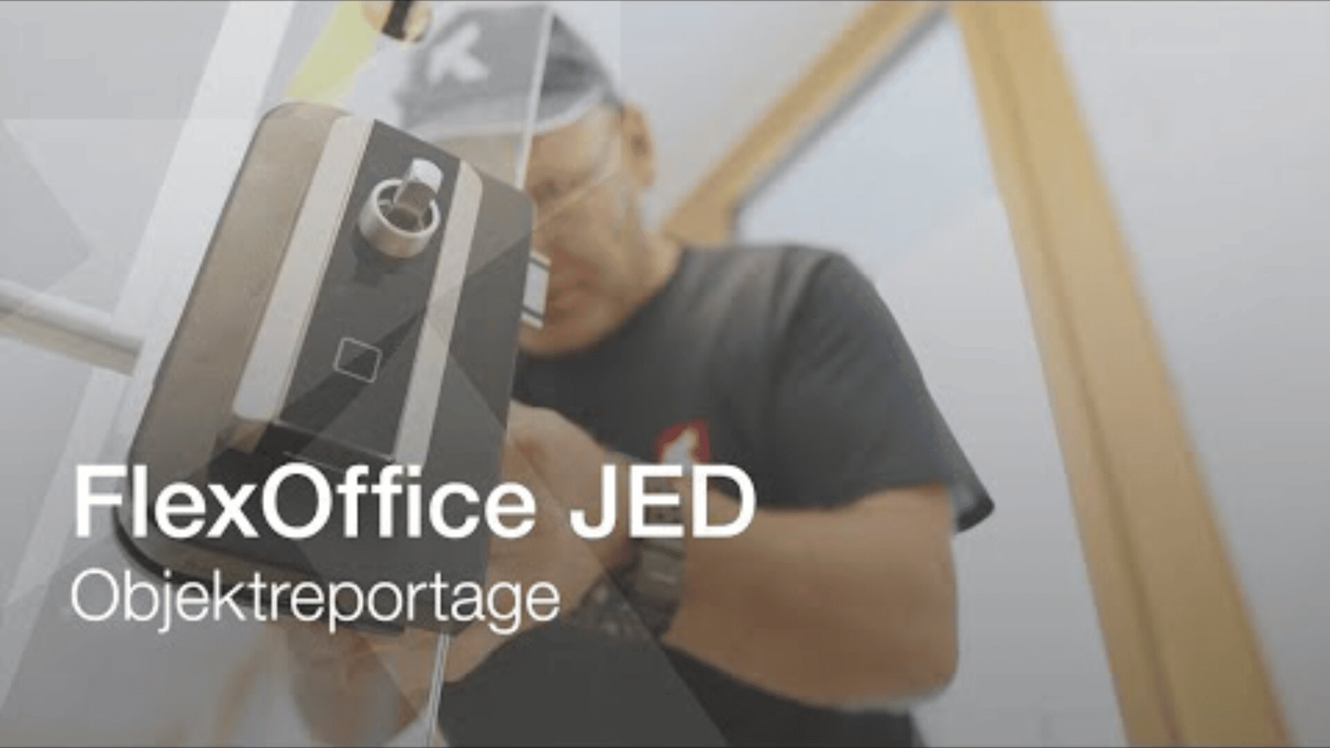Eine Person installiert oder repariert ein Schloss an einer Glastür. Der überlagerte Text lautet FlexOffice JED Objektreportage. Das Bild ist leicht unscharf und scheint Teil eines Berichts oder einer Fallstudie zu sein.