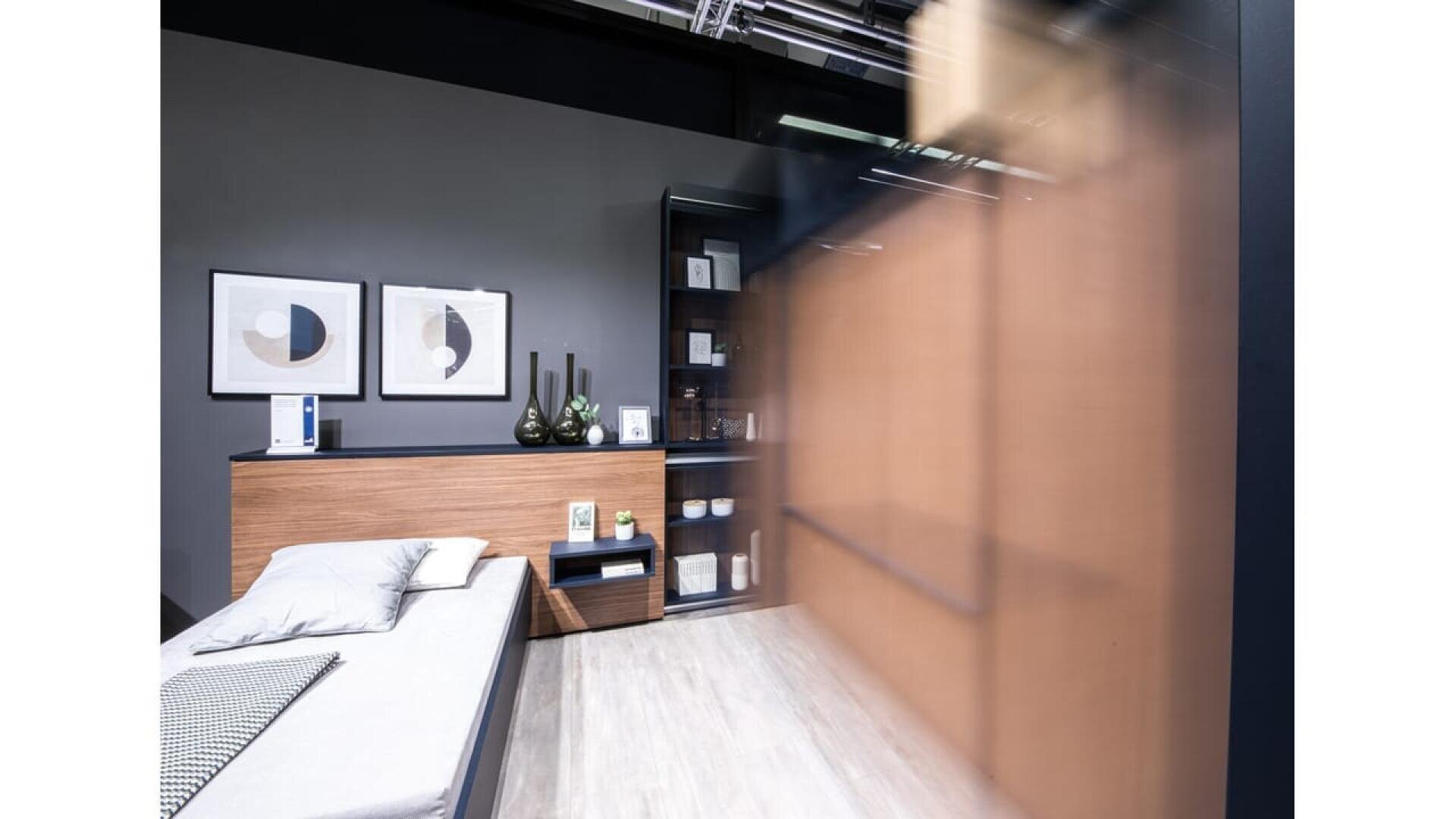 Ein modernes, minimalistisches Schlafzimmer mit einer grauen Wand, einem Bett mit weißer Bettwäsche, Holzmöbeln, abstrakter Kunst, Regalen mit Dekoration und einer verschwommenen Holzschiebetür.