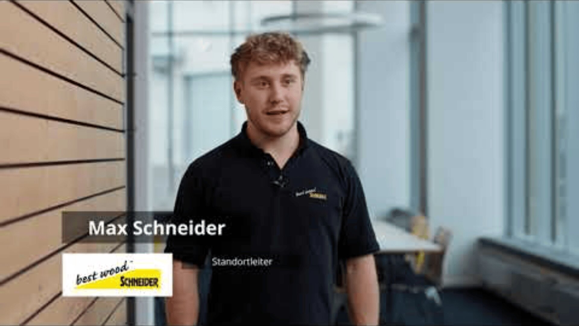 Ein Mann in einem schwarzen SCHNEIDER-Hemd aus bestem Holz steht in einem Innenraum an einer Holzwand. Der Text auf dem Bild lautet Max Schneider, Standortleiter, darunter das SCHNEIDER-Logo für best wood.