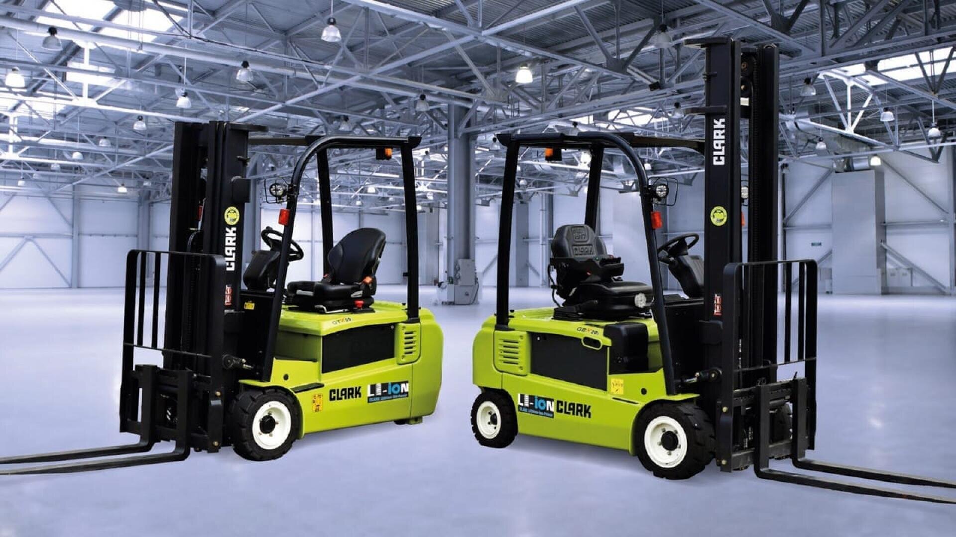 Deux chariots élévateurs verts CLARK Li-Ion sont garés à l'intérieur d'un entrepôt spacieux et bien éclairé, avec un plafond élevé et des poutres métalliques. Les chariots se font face et l'entrepôt semble vide.
