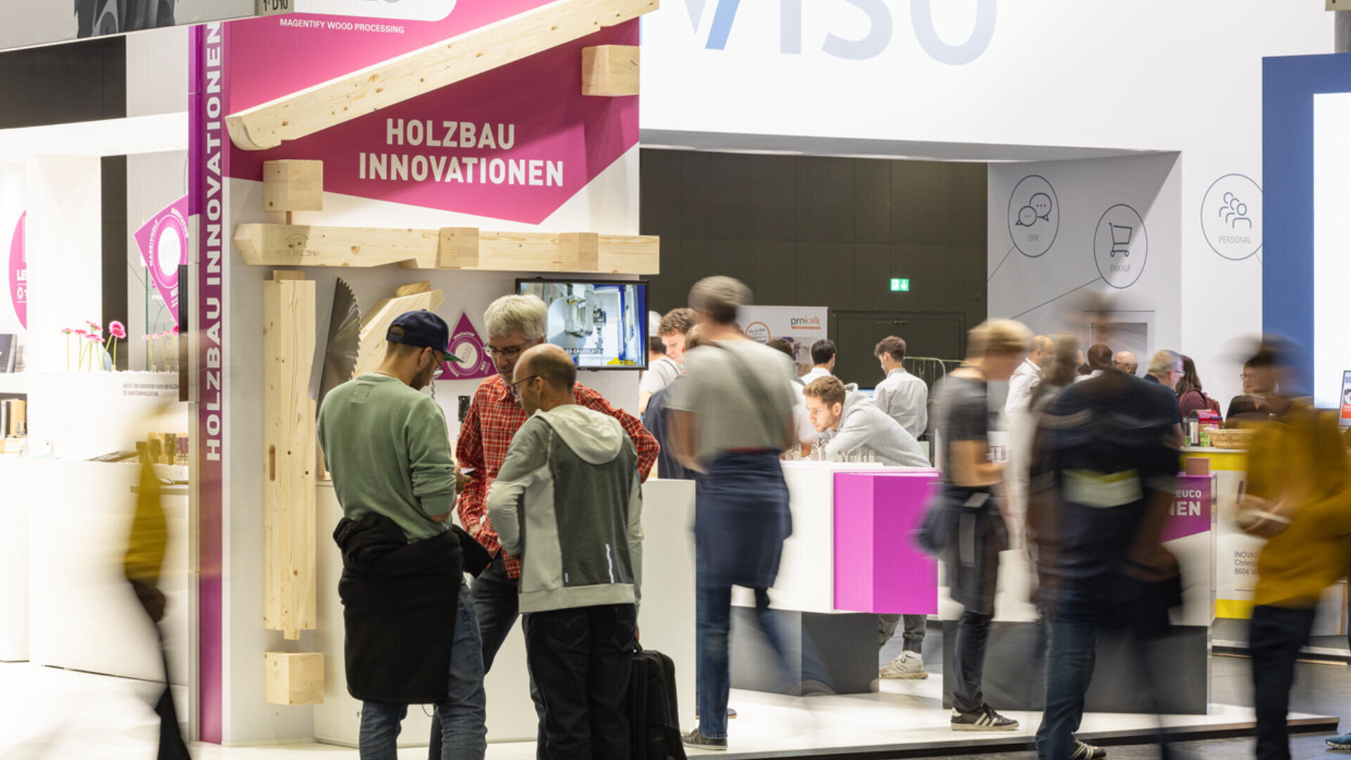 Menschen versammeln sich an einem Messestand mit der Aufschrift "HOLZBAU INNOVATIONEN" und einer leuchtend rosa Beschriftung. Der Stand besteht aus einer Holzstruktur und einem Bildschirm, an dem die Besucher in einer belebten Halle vorbeilaufen und sich unterhalten.