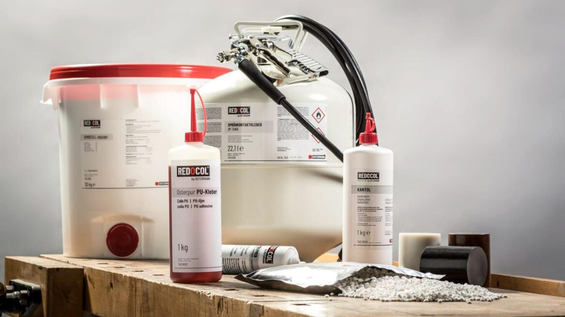 Mehrere Behälter unterschiedlicher Größe, die mit industriellen RED COOL Produkten gefüllt sind, darunter Flaschen, ein Fass und Säcke, sind auf einer Holzfläche mit einigen Pellets und zylindrischen festen Proben angeordnet.
