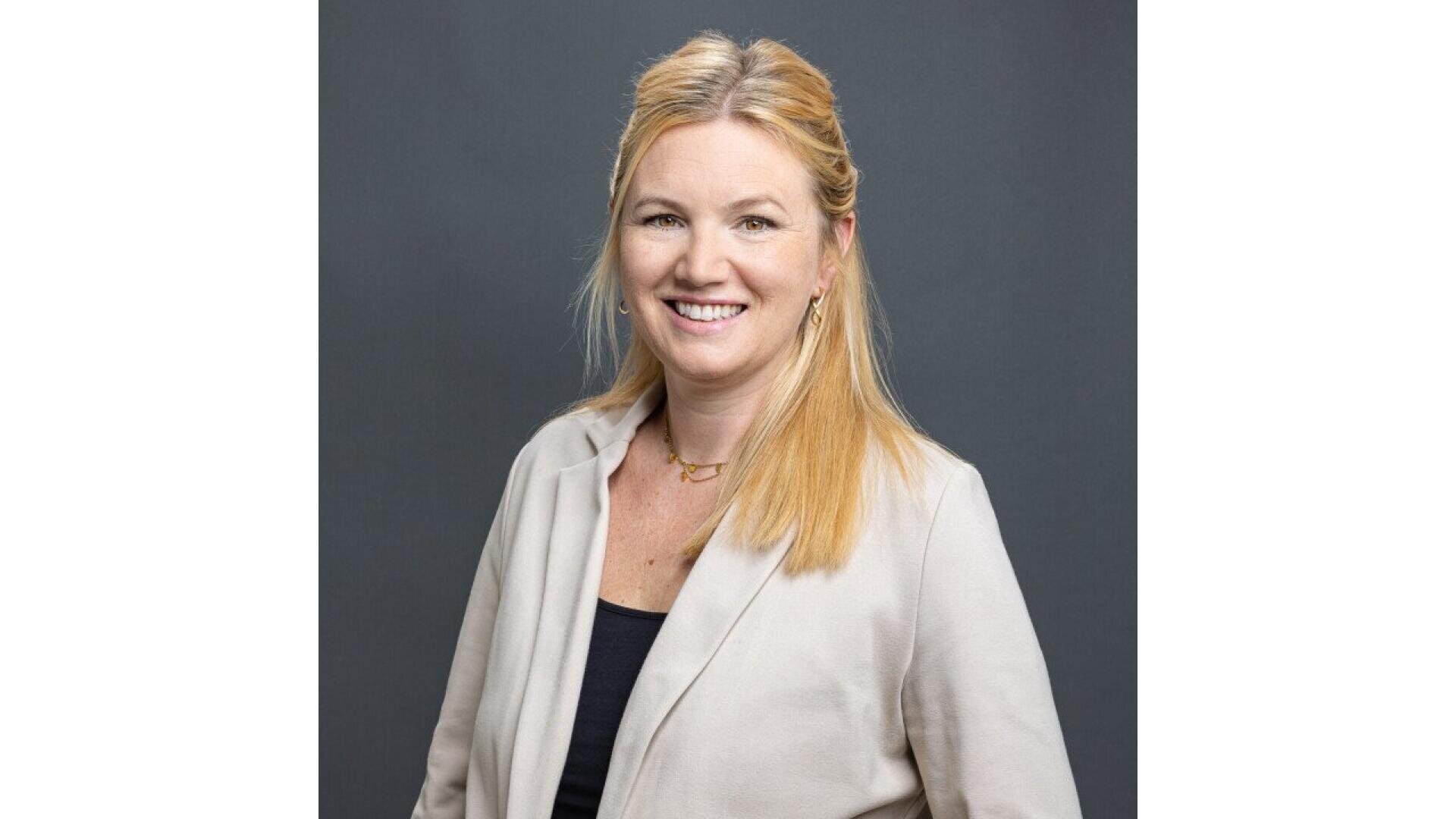 Une femme souriante aux longs cheveux blonds, portant un blazer beige clair sur un haut sombre, se tient devant un fond gris uni.