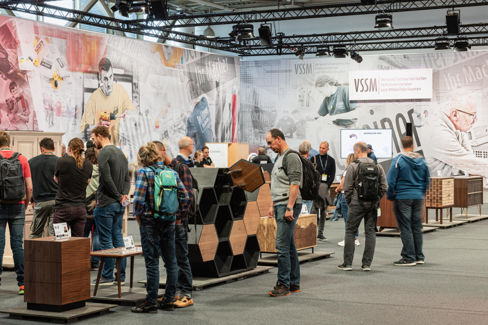 Un groupe de personnes visite un stand de salon professionnel présentant des meubles et des présentoirs en bois. Le stand comporte des panneaux d'information et une grande fresque d'images de travail du bois sur le mur du fond. Certains participants interagissent avec les objets exposés.