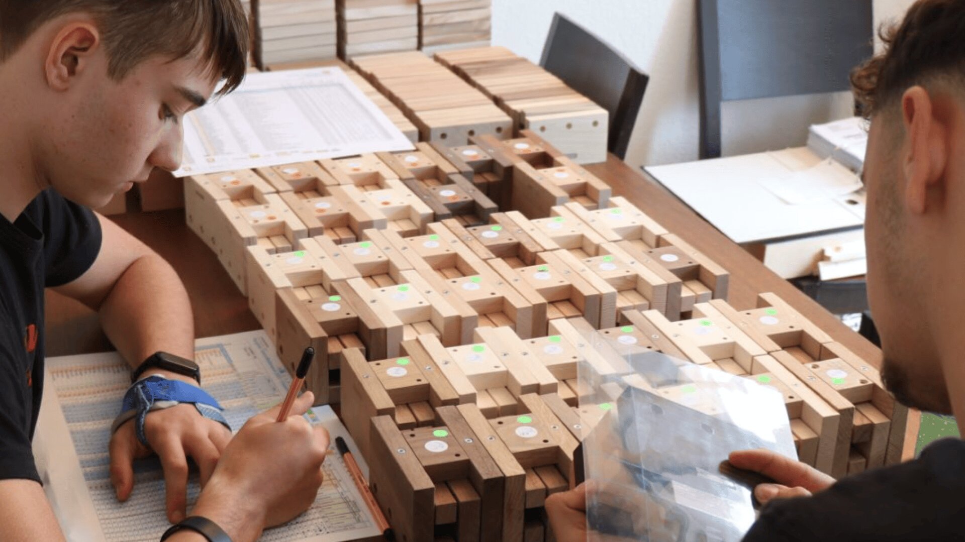 Deux personnes sont assises à une table avec des blocs de bois, chacun marqué avec des autocollants de couleur. Une personne écrit sur une fiche de données, tandis qu'une autre manipule un sac. D'autres blocs de bois et d'autres papiers sont étalés sur la table.