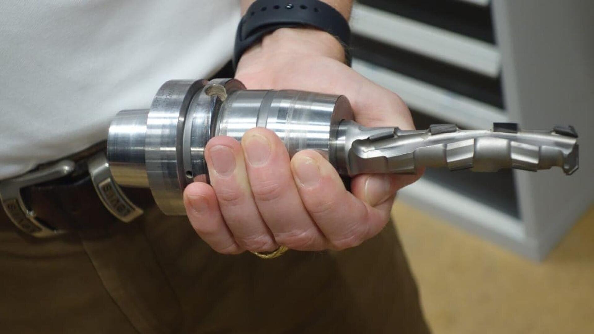 Eine Person mit weißem Hemd und brauner Hose hält ein großes, metallisches Industriewerkzeug oder Maschinenteil mit zylindrischer Basis und spiralförmiger Schneide.