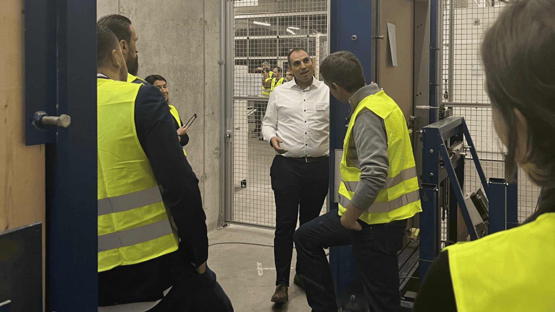 Un groupe de personnes portant des gilets de sécurité jaunes écoute un homme en chemise blanche qui parle dans un environnement industriel avec des murs en béton et des clôtures métalliques.