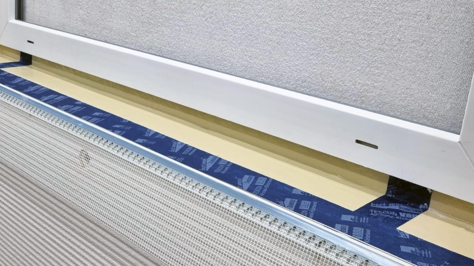 Nahaufnahme eines Baudetails, das einen weißen Fensterrahmen über einem gelben Streifen, blaues Schutzband und ein an einer Wand befestigtes Metallgitter zeigt, das wahrscheinlich Teil eines Isolier- oder Veredelungsprozesses ist.