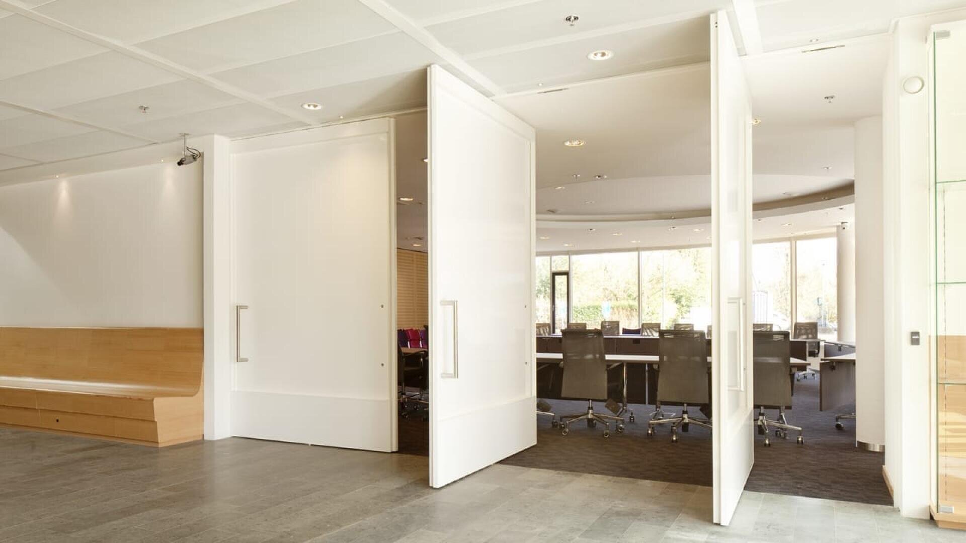 Espace de bureau moderne avec de grandes portes blanches à double battant qui s'ouvrent pour révéler une salle de conférence avec plusieurs chaises autour d'une longue table. L'espace est bien éclairé par la lumière naturelle provenant de hautes fenêtres.
