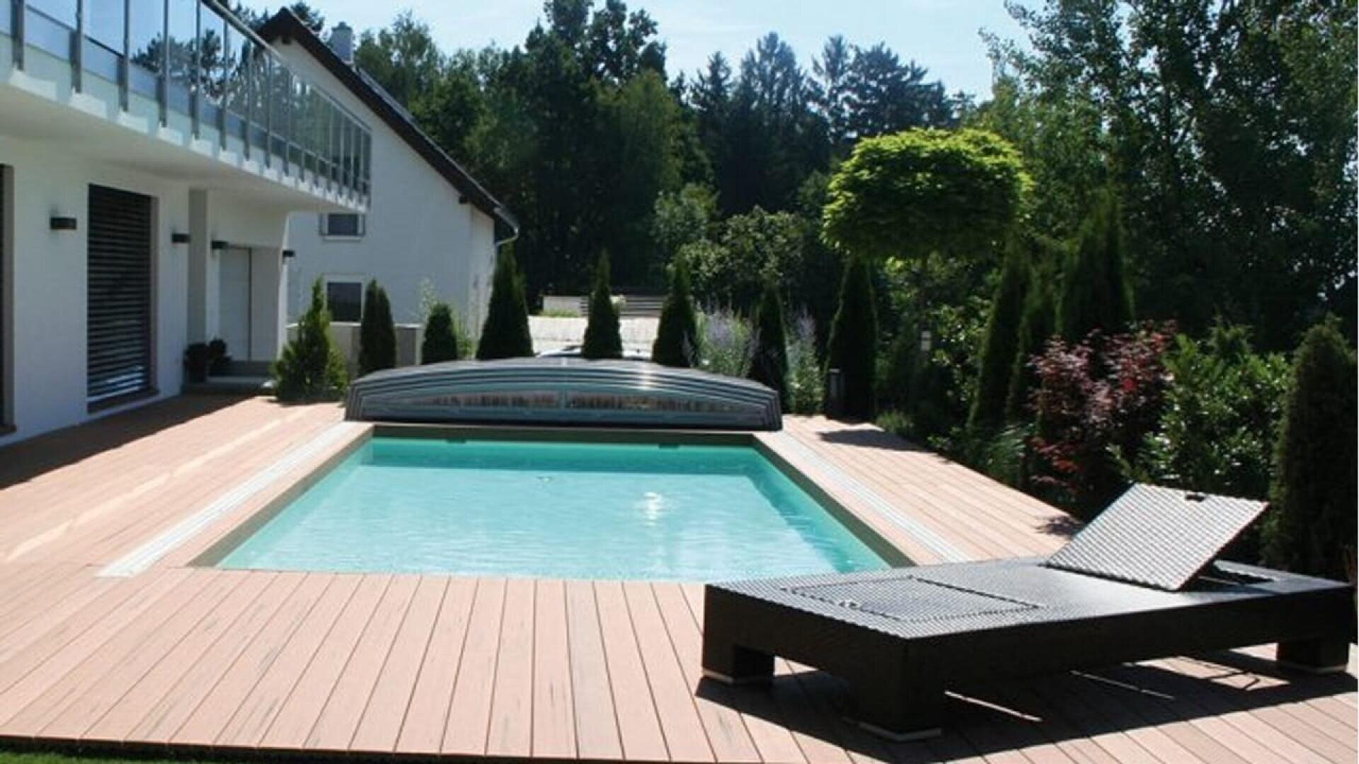 Une piscine extérieure rectangulaire à l'eau claire est entourée d'une terrasse en bois. Une chaise longue en osier noir est installée au bord de la piscine. Des arbres et des arbustes se trouvent dans le jardin paysager, à côté d'un bâtiment moderne.
