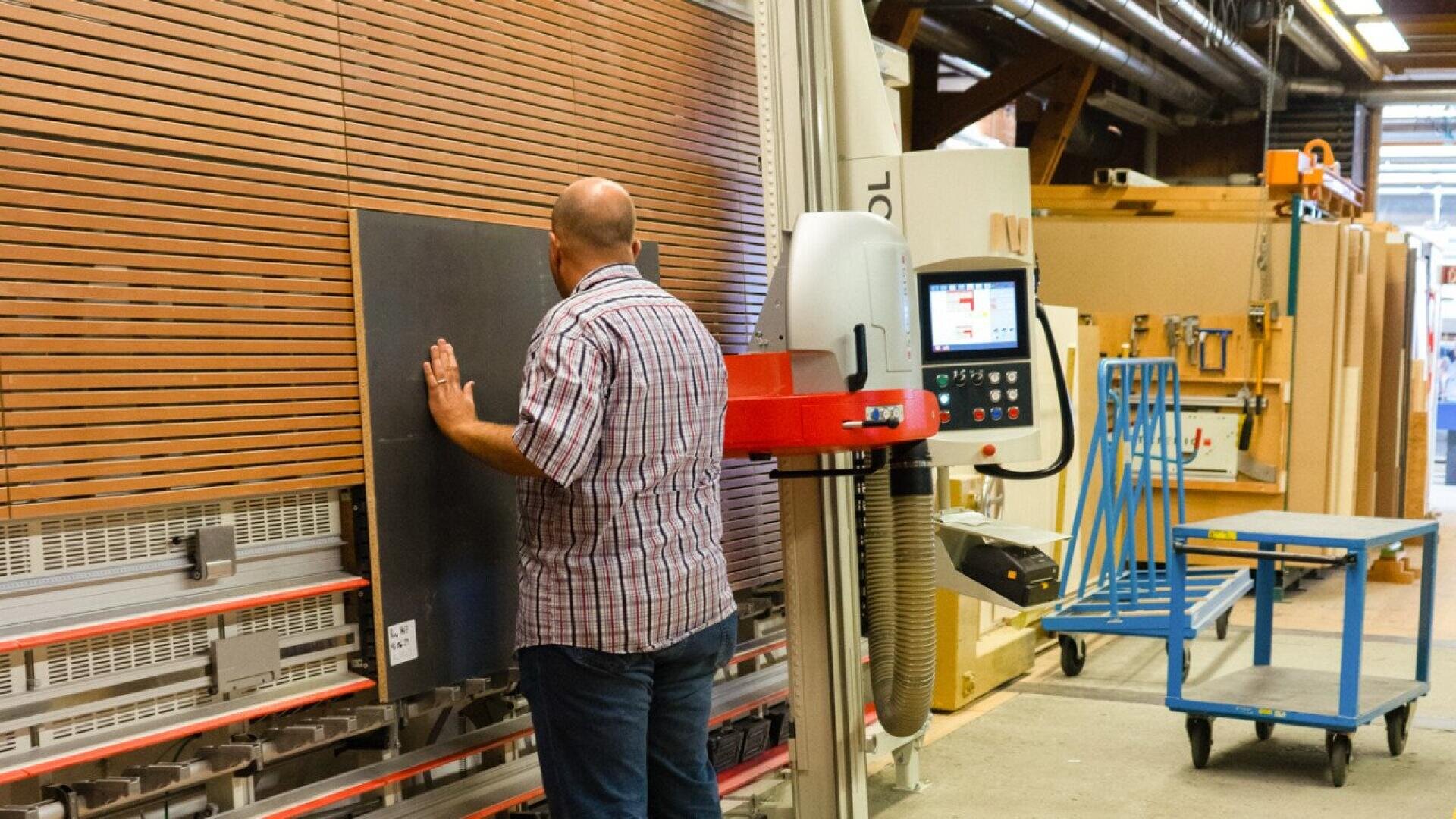 Un homme en chemise à carreaux utilise une grande scie verticale dans un atelier de menuiserie, positionnant un panneau sombre pour le couper. Des équipements et des outils industriels sont visibles à l'arrière-plan.