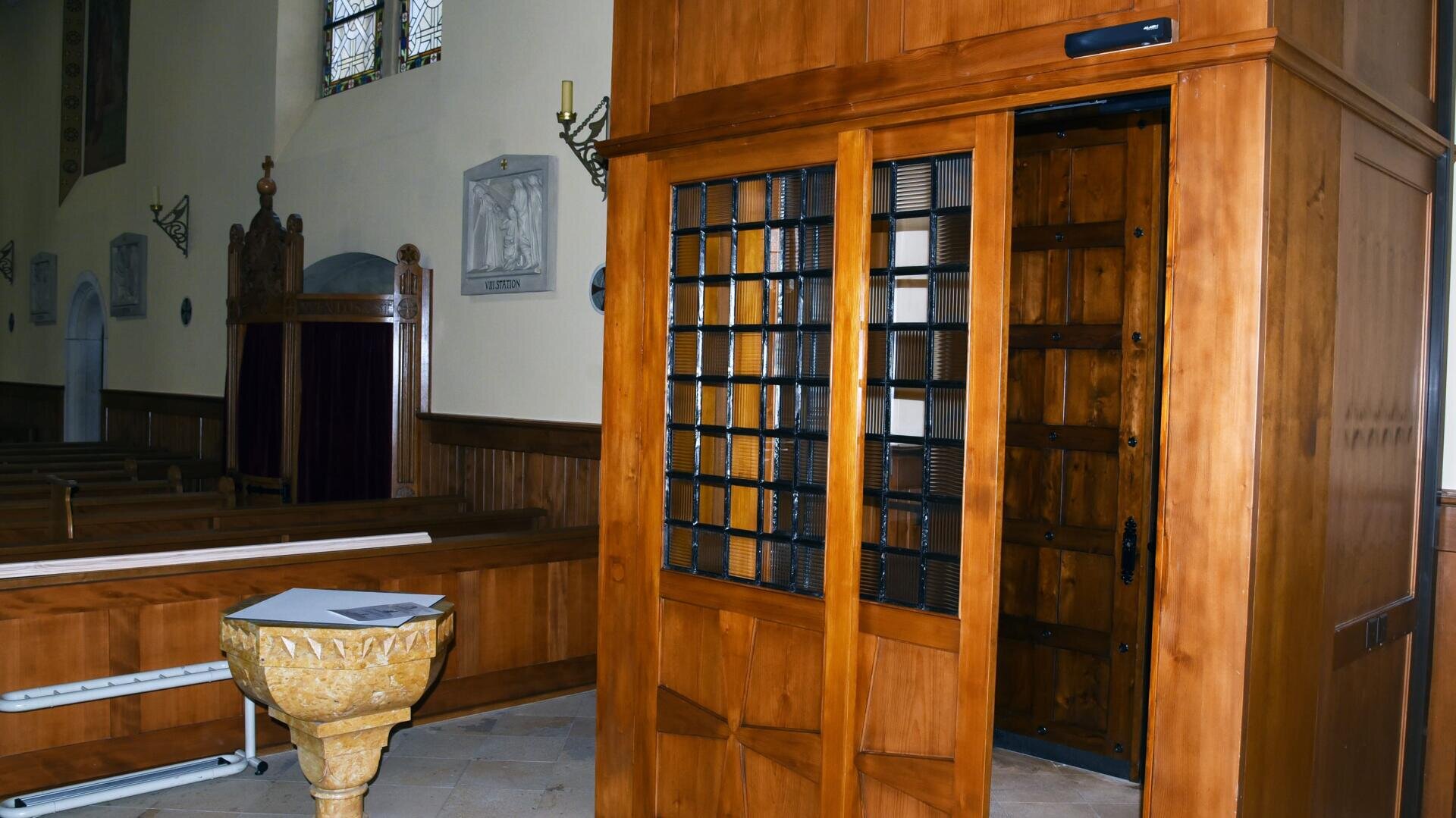 Un confessionnal en bois avec une fenêtre grillagée se trouve à l'intérieur d'une église, près de bancs en bois et de fonts baptismaux. Des vitraux et des plaques religieuses sont visibles sur les murs.