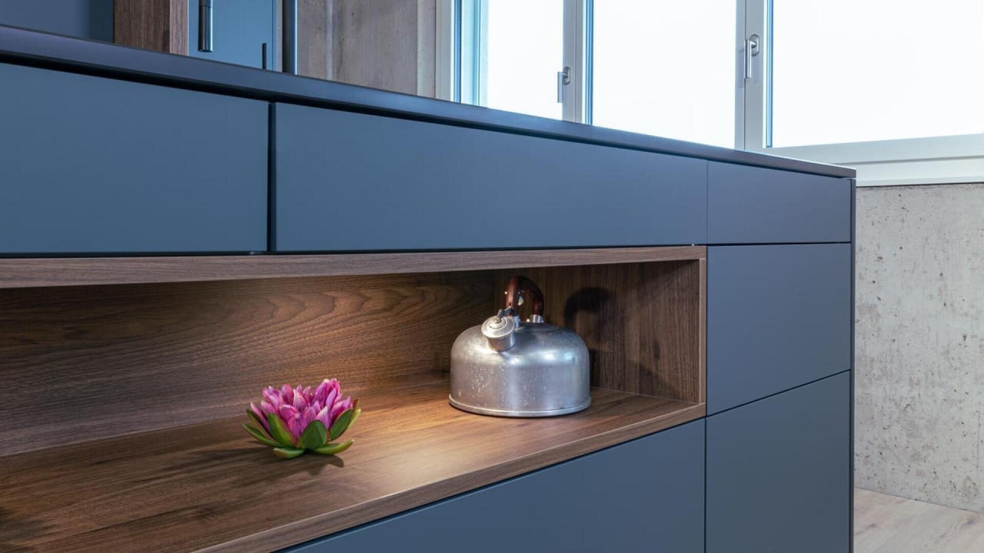Un meuble de cuisine moderne avec des tiroirs bleu-gris mats et une étagère ouverte en bois, présentant une bouilloire argentée et une fleur rose. De grandes fenêtres à l'arrière-plan laissent entrer la lumière naturelle.