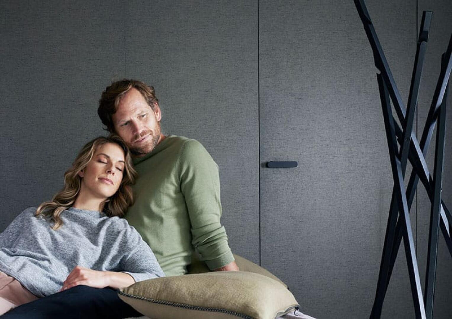 Eine Frau mit geschlossenen Augen legt ihren Kopf auf die Schulter eines Mannes, während sie eng beieinander auf einem Sofa sitzen. Beide wirken entspannt und ruhig in einem modernen, minimalistischen Raum mit einer grauen Wand und abstraktem schwarzen Dekor.