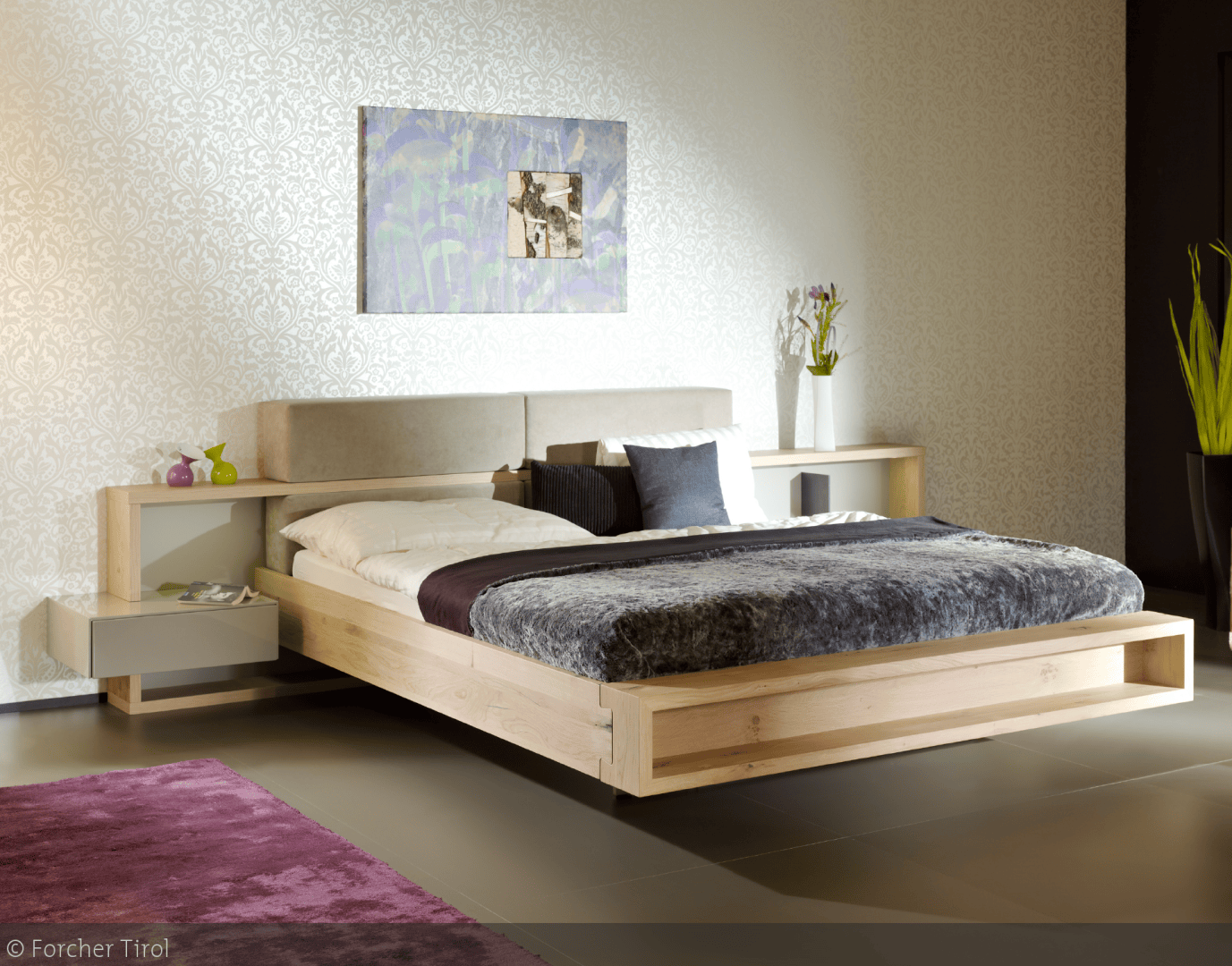 Ein modernes Schlafzimmer mit einem hellen Holzpritschenbett, grauer und weißer Bettwäsche, wandmontierten Nachttischen, Blumentapete, einem lila Teppich und einem Gemälde über dem Bett. Vasen und Dekoration stehen auf den Regalen am Kopfende des Bettes.