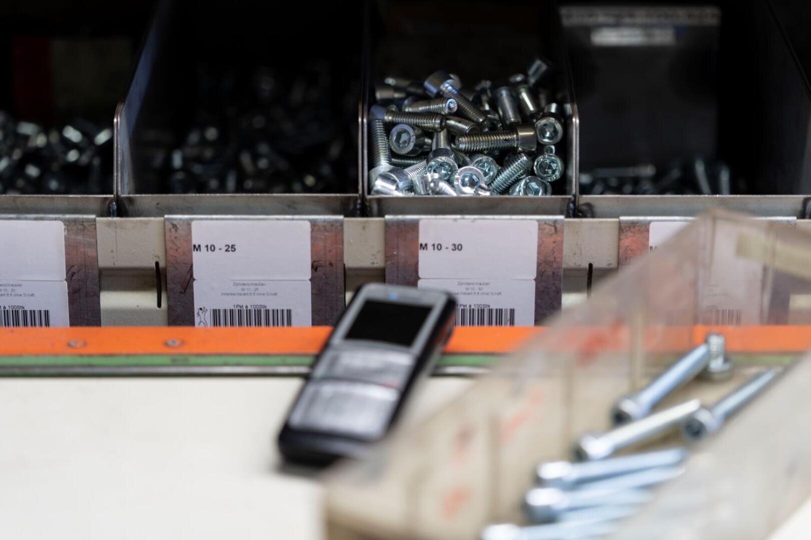 Nahaufnahme von Metallbehältern mit der Aufschrift "M 10 - 25" und "M 10 - 30", die mit Schrauben und Bolzen gefüllt sind. Ein mobiles Gerät steht vorne auf dem Tresen und weitere Schrauben sind im Vordergrund zu sehen.