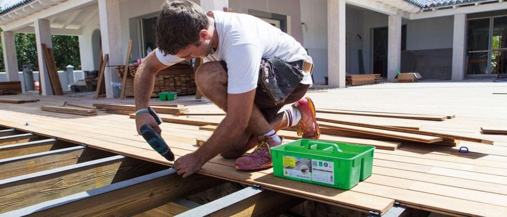 Ein Mann geht in die Hocke, während er mit einer Bohrmaschine Holzbretter an einem Rahmen für eine Außenterrasse befestigt. Neben ihm steht ein grüner Werkzeugkasten, und im Hintergrund ist ein Haus zu sehen.