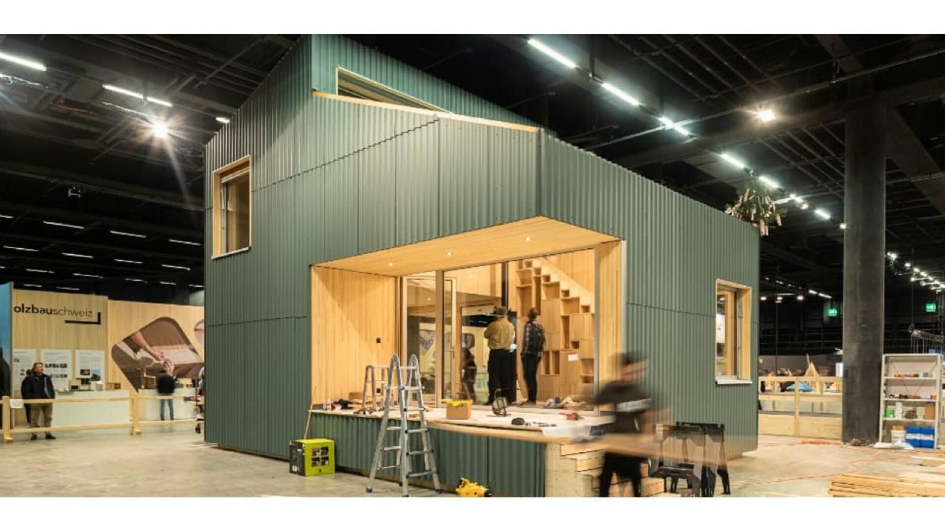 Une petite maison verte moderne avec de grandes fenêtres est présentée à l'intérieur lors d'une exposition. Plusieurs personnes s'installent ou travaillent autour d'elle, des outils et une échelle sont visibles, et l'intérieur révèle des escaliers en bois et un éclairage chaleureux.