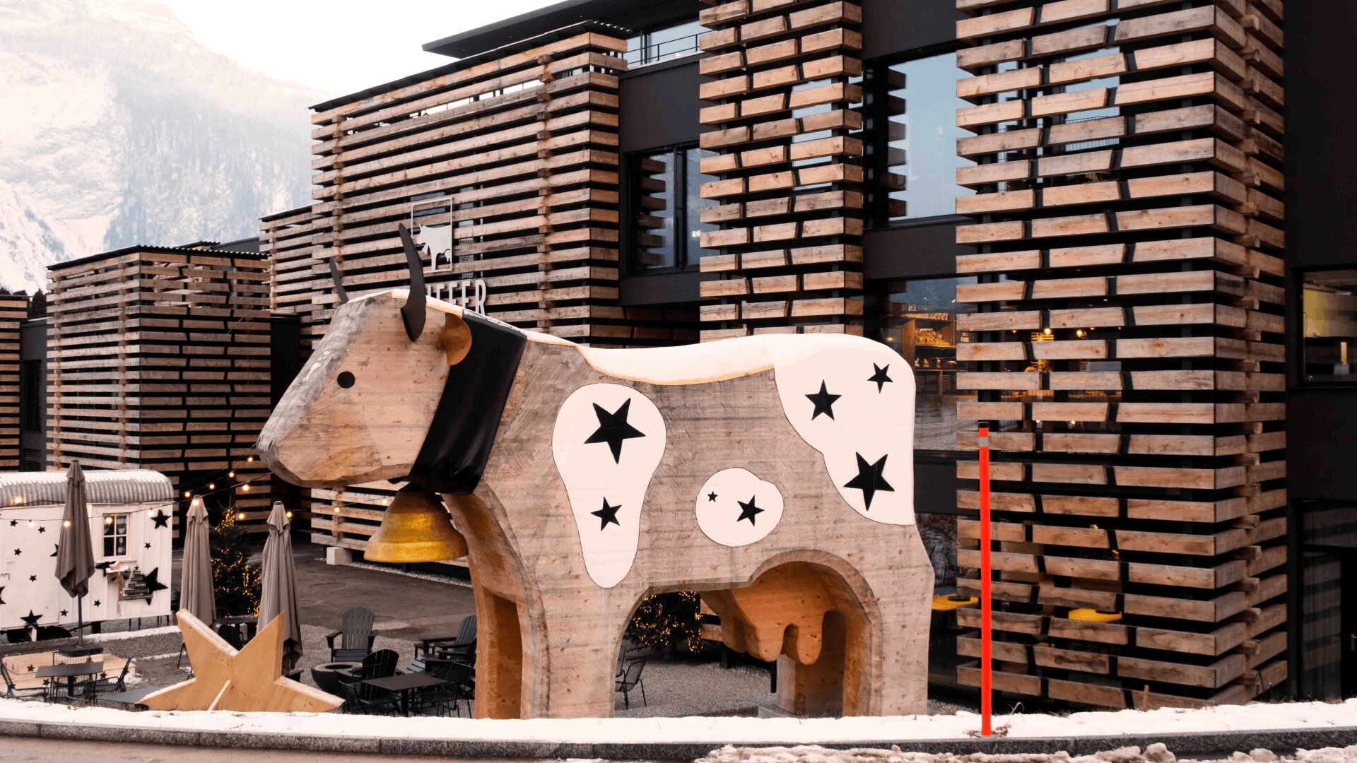 Une grande sculpture de vache en bois avec des étoiles noires et une écharpe se trouve devant un bâtiment moderne avec des panneaux de bois à lattes horizontales. La neige et les montagnes sont visibles à l'arrière-plan.