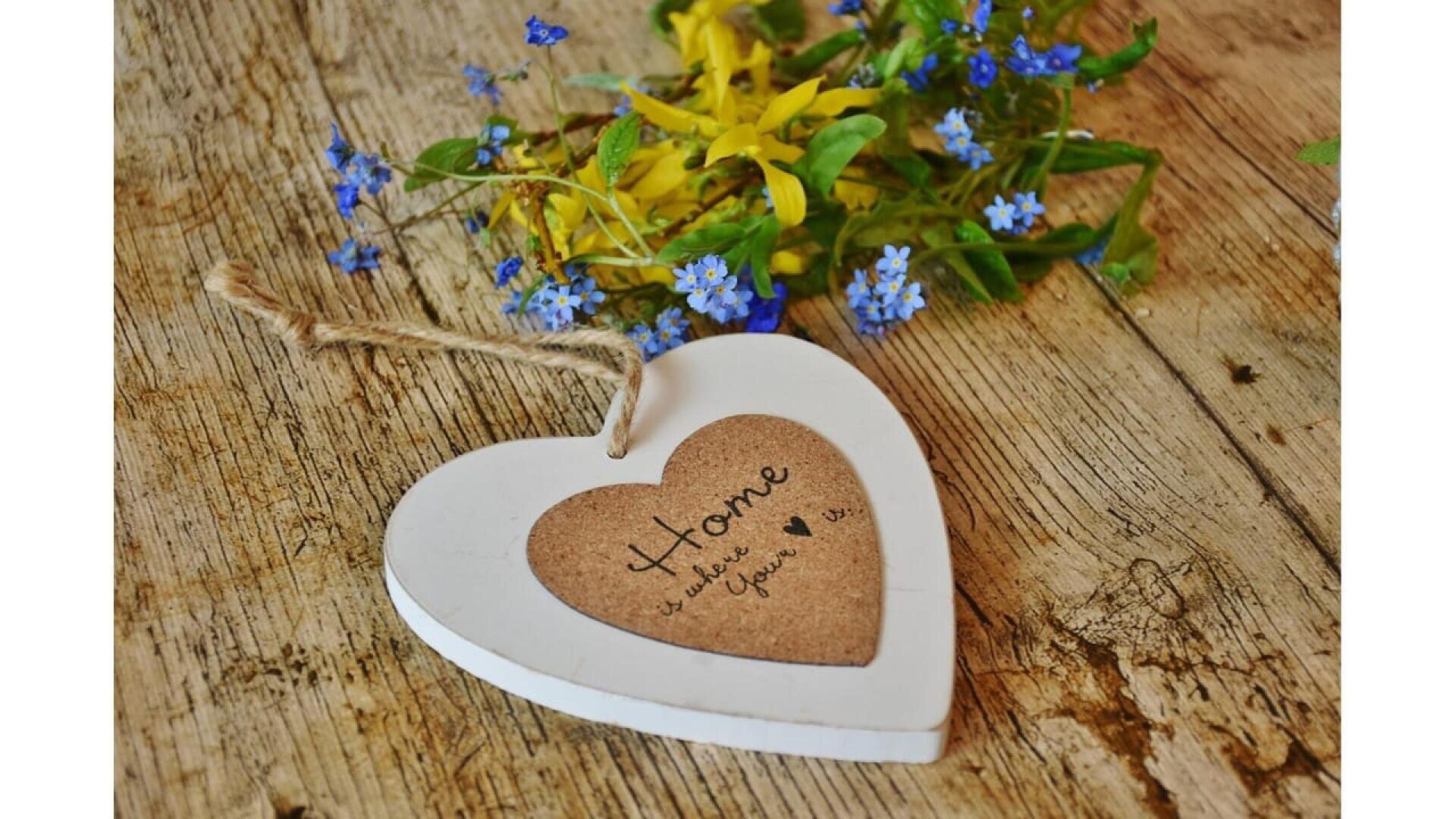 Un panneau en bois en forme de cœur sur lequel est écrit Home is where the heart est posé sur une surface rustique en bois, à côté de petites fleurs jaunes et bleues.