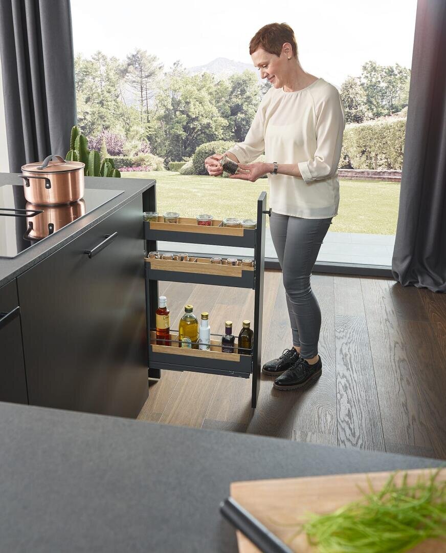 Eine Frau steht in einer modernen Küche neben einem ausziehbaren Gewürzregal und hält ein Gewürzglas in der Hand. In dem Regal sind Flaschen mit Öl und Gewürzen aufgereiht. Große Fenster hinter ihr geben den Blick auf eine grüne, malerische Landschaft frei.