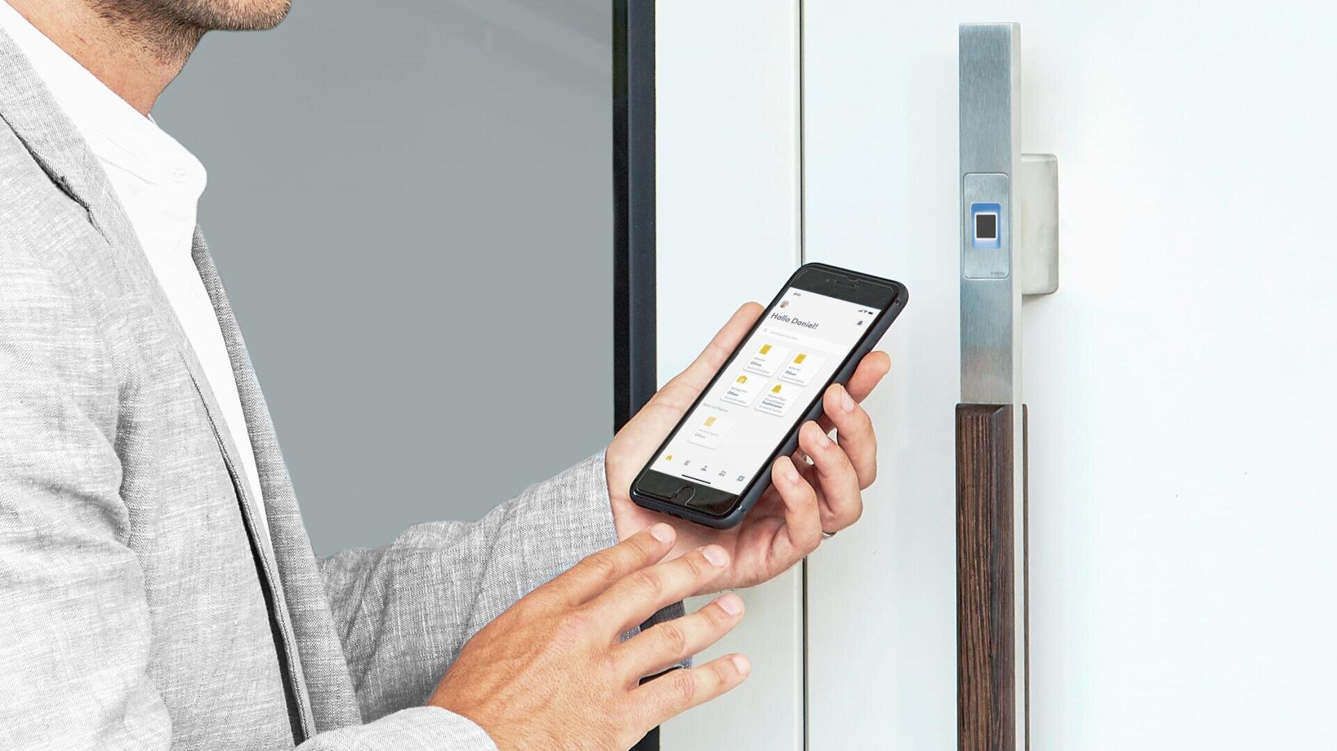 Une personne en costume gris clair tient un smartphone affichant une application de serrure intelligente devant une porte moderne équipée d'une serrure électronique.