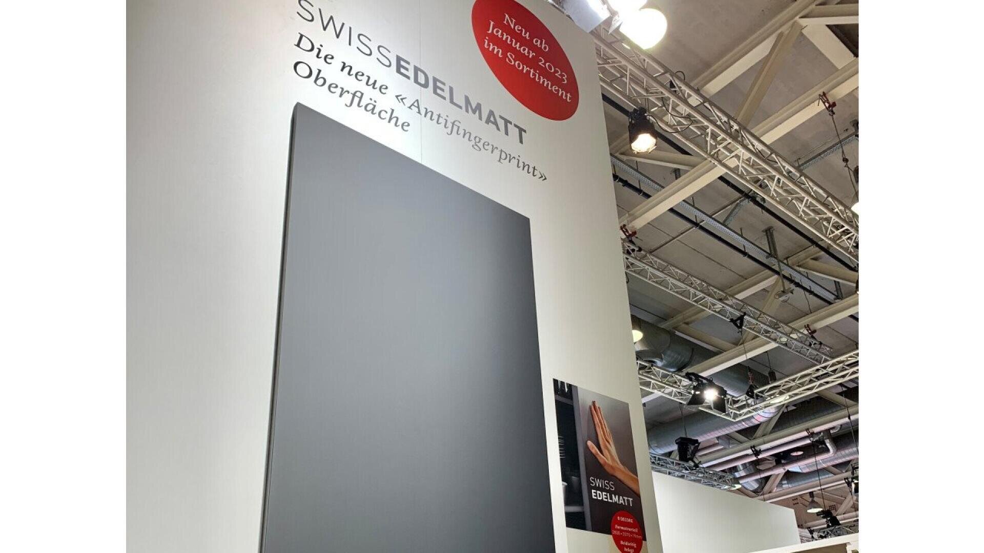 Sur un mur d'exposition, un grand panneau sombre présente la nouvelle surface anti-traces de doigts SWISSEDELAMATT, avec un texte en allemand et des lumières au plafond visibles au-dessus.