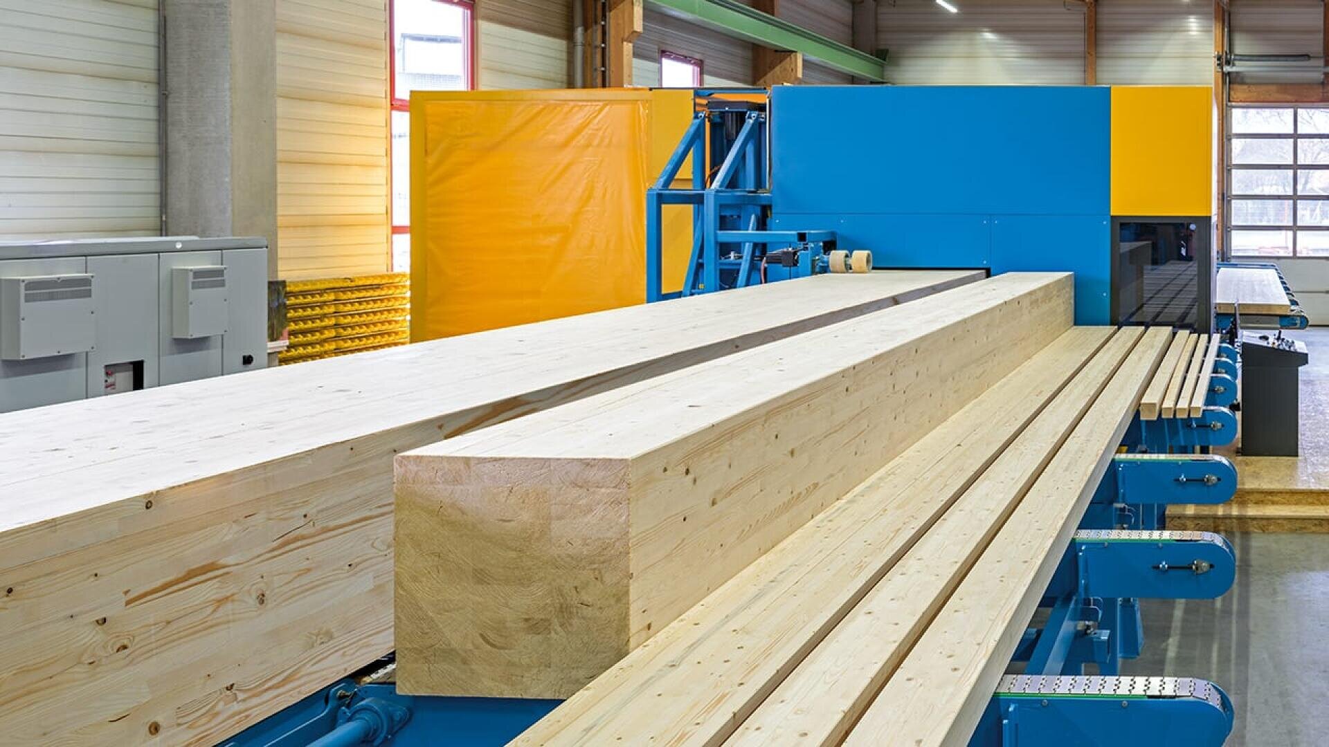 Große Brettschichtholzbalken und -bretter auf einem Fördersystem in einem modernen holzverarbeitenden Betrieb mit industrieller Ausstattung und heller Innenbeleuchtung.
