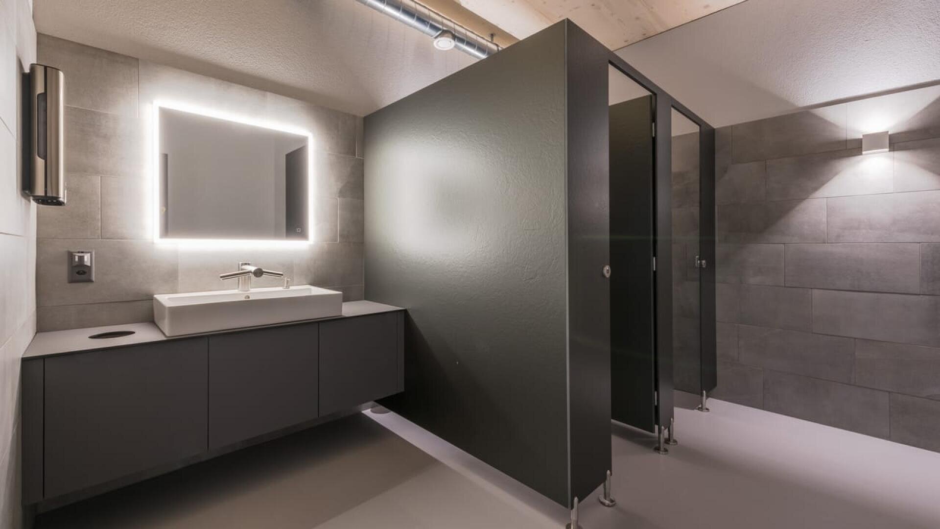 Toilettes publiques modernes avec un miroir rétroéclairé au-dessus d'un lavabo et d'un comptoir, et deux cabines de toilettes fermées avec des cloisons sombres. L'espace est doté d'un carrelage gris et d'un éclairage zénithal doux.