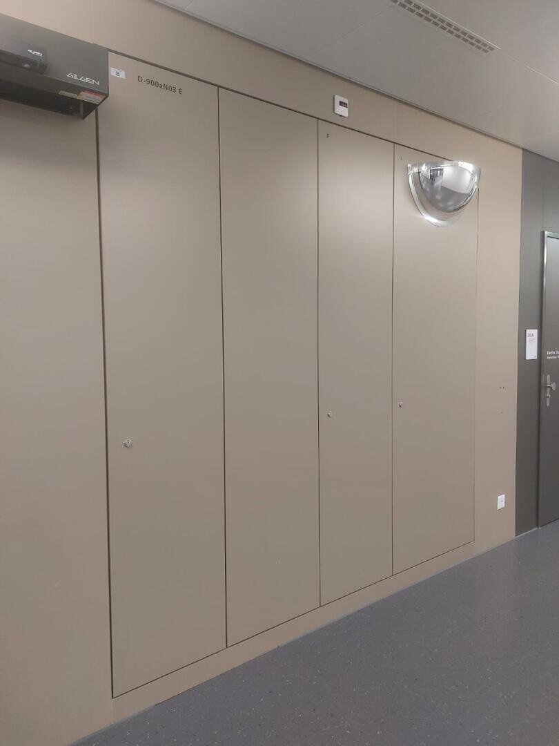 Minimalistische beige Wand mit vier hohen, schmalen, bündigen Türen; rechts befindet sich eine kuppelförmige Wandleuchte. Der Boden ist dunkelgrau, und in der oberen linken Ecke ist ein Teil eines schwarzen Geräts zu sehen.