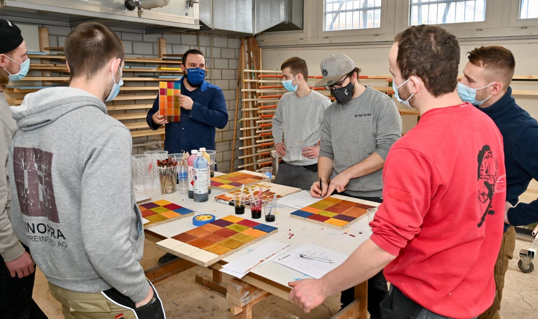 Eine Gruppe von sechs Personen mit Gesichtsmasken versammelt sich um einen Tisch mit bunten Holzfliesen, Bechern mit Flüssigkeit und Papieren, während eine Person etwas erklärt und eine Musterfliese in einer Werkstattumgebung hochhält.