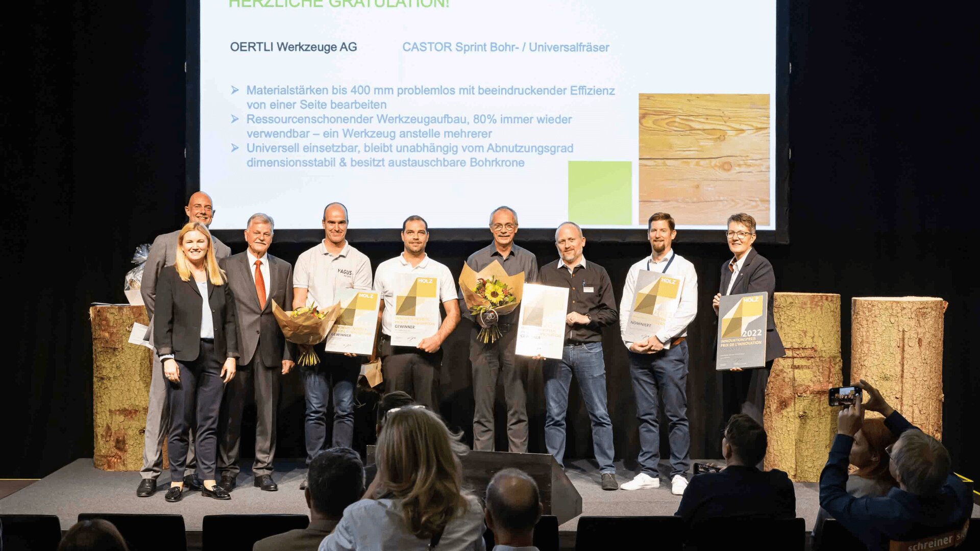 Eine Gruppe von zehn Personen steht auf der Bühne, hält Urkunden und Blumen in der Hand und posiert für ein Foto vor einer Präsentationswand mit Unternehmensinformationen. Im Vordergrund ist ein Publikum zu sehen.