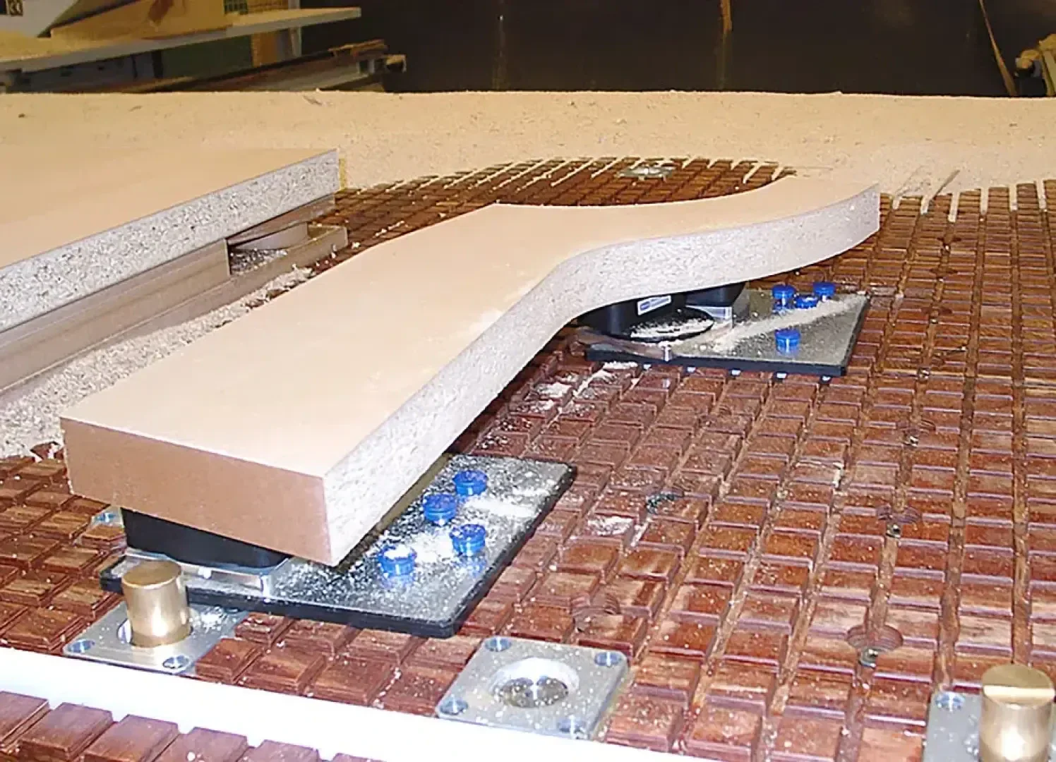 Eine gebogene Holzplatte liegt auf einem CNC-Maschinentisch, der von Vakuumkapseln gehalten wird, und auf dem Holzstaub von der letzten Bearbeitung verstreut ist.