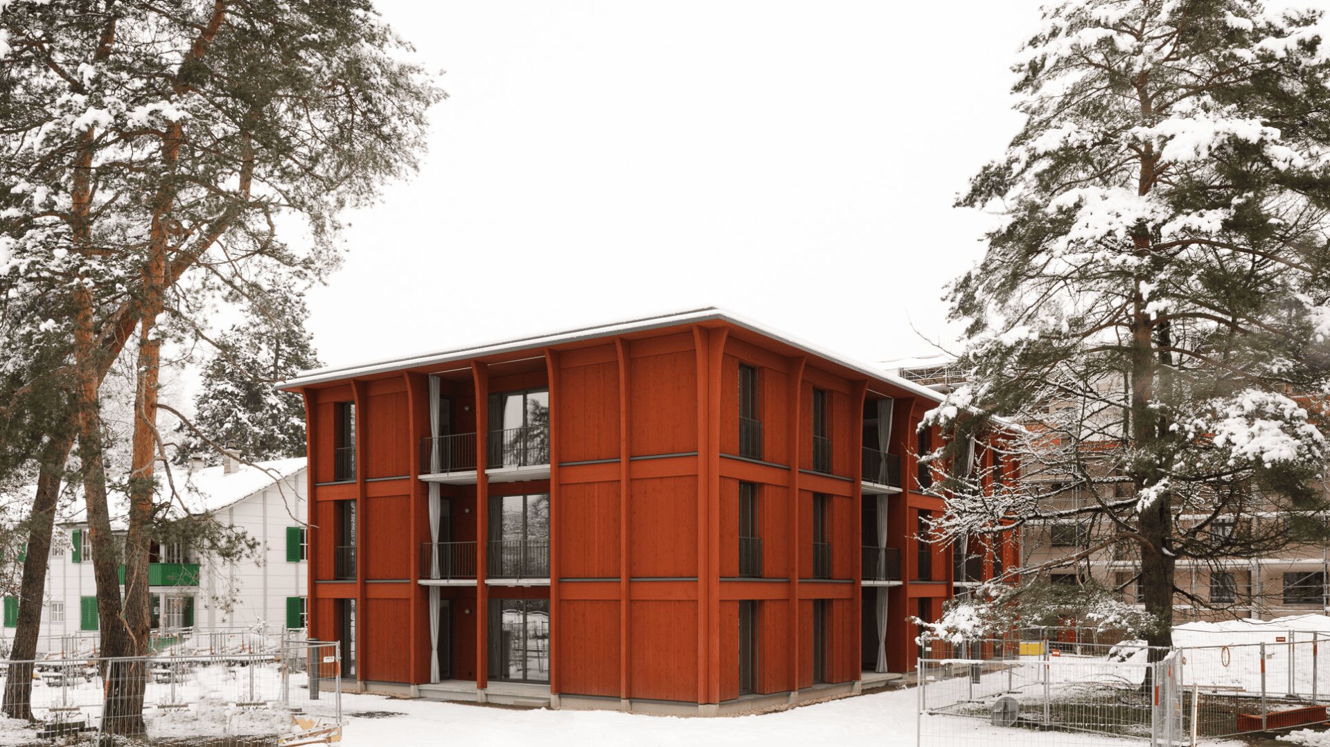 Ein modernes, rotes, vierstöckiges Gebäude mit großen Fenstern ist von schneebedeckten Bäumen und dem Boden umgeben, eingebettet in eine winterliche Landschaft mit nahen weißen Gebäuden und einem Drahtzaun im Vordergrund.