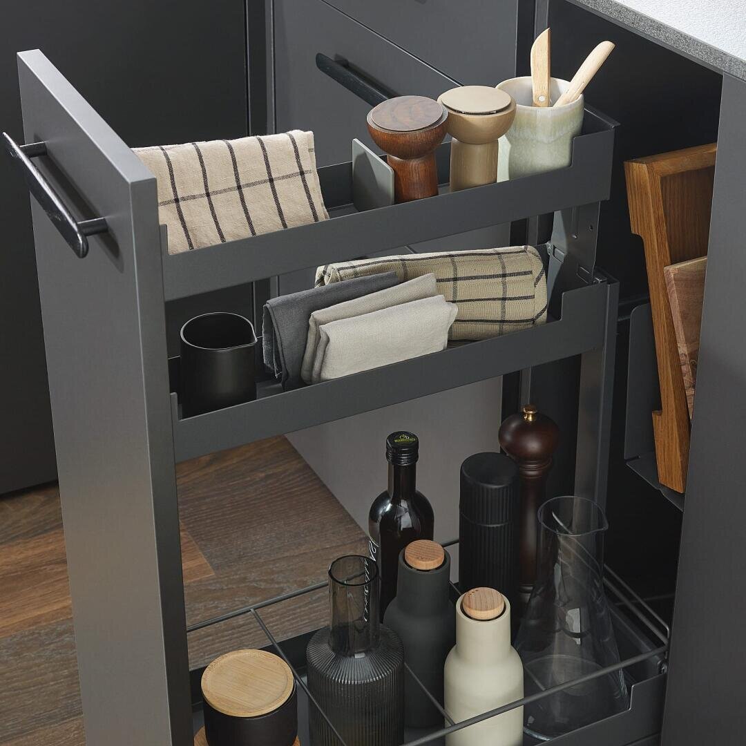 Ein ausziehbarer Küchenschrank mit Regalen mit Flaschen, Gläsern, gefalteten Geschirrtüchern, Holzutensilien, Pfeffermühlen und Glasbehältern, die fein säuberlich vor einem dunklen Holzboden angeordnet sind.