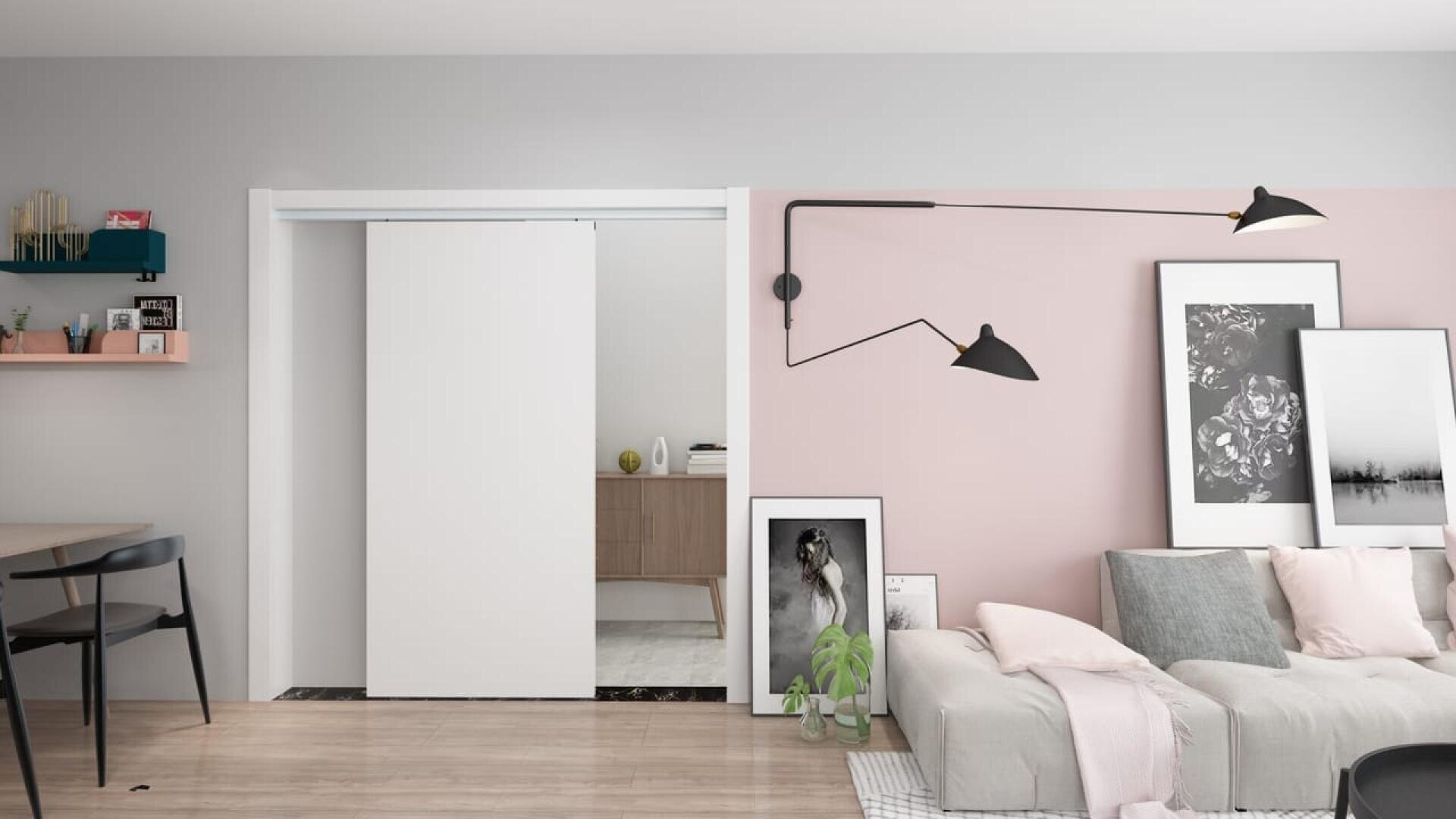 Salon moderne avec des planchers en bois clair, un canapé gris, des œuvres d'art encadrées sur un mur rose chair, une lampe murale noire et une porte coulissante blanche menant à une autre pièce. Une petite plante est posée près du canapé.