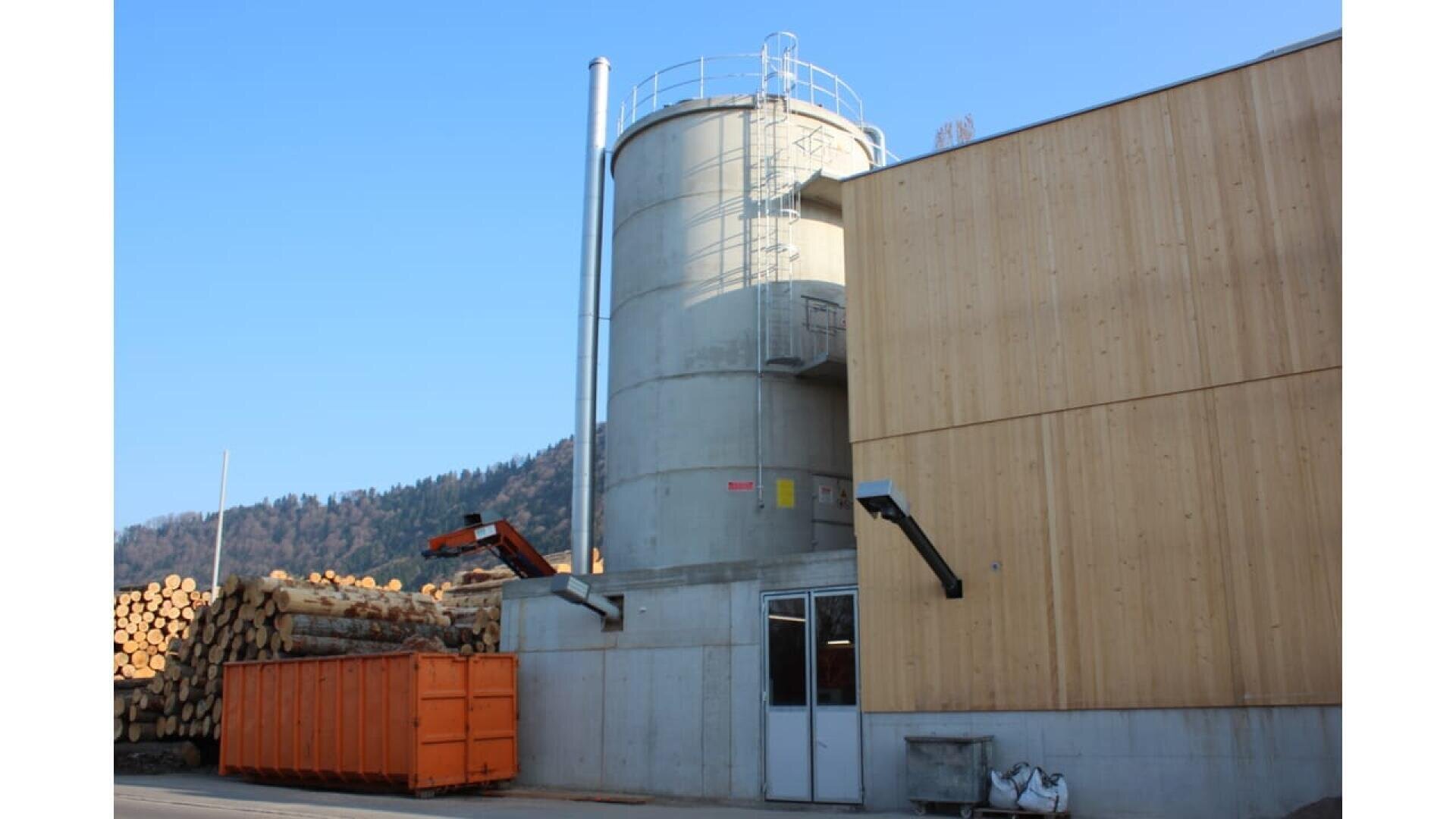 Un haut silo cylindrique en béton avec un escalier en métal est attaché à un bâtiment en bois. Des grumes empilées et une benne à ordures orange sont visibles à proximité, avec des collines et un ciel bleu clair en arrière-plan.
