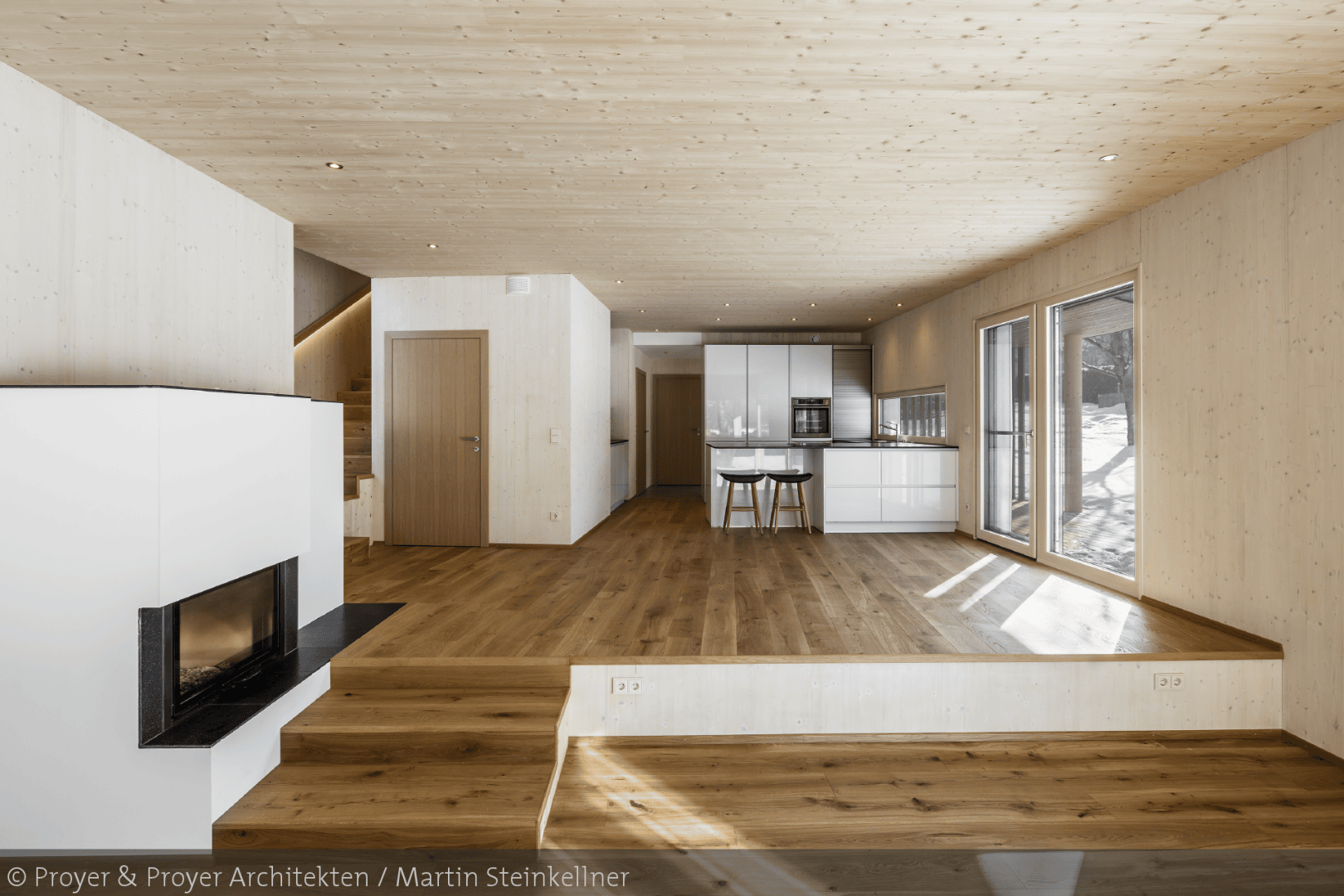 Modernes, offenes Wohnkonzept mit Holzböden, -decken und -wänden, weißem Kamin, Küche mit weißen Schränken und schwarzen Arbeitsplatten, Esstisch, großen Fenstern und natürlichem Licht, das hereinströmt. Durch die Fenster ist der Schnee draußen sichtbar.