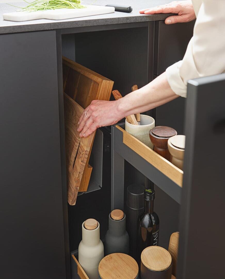 Eine Person bewahrt ein hölzernes Schneidebrett in einem dunklen Küchenschrank auf, in dem verschiedene Flaschen, Gläser und Küchenutensilien übersichtlich auf Regalen angeordnet sind.