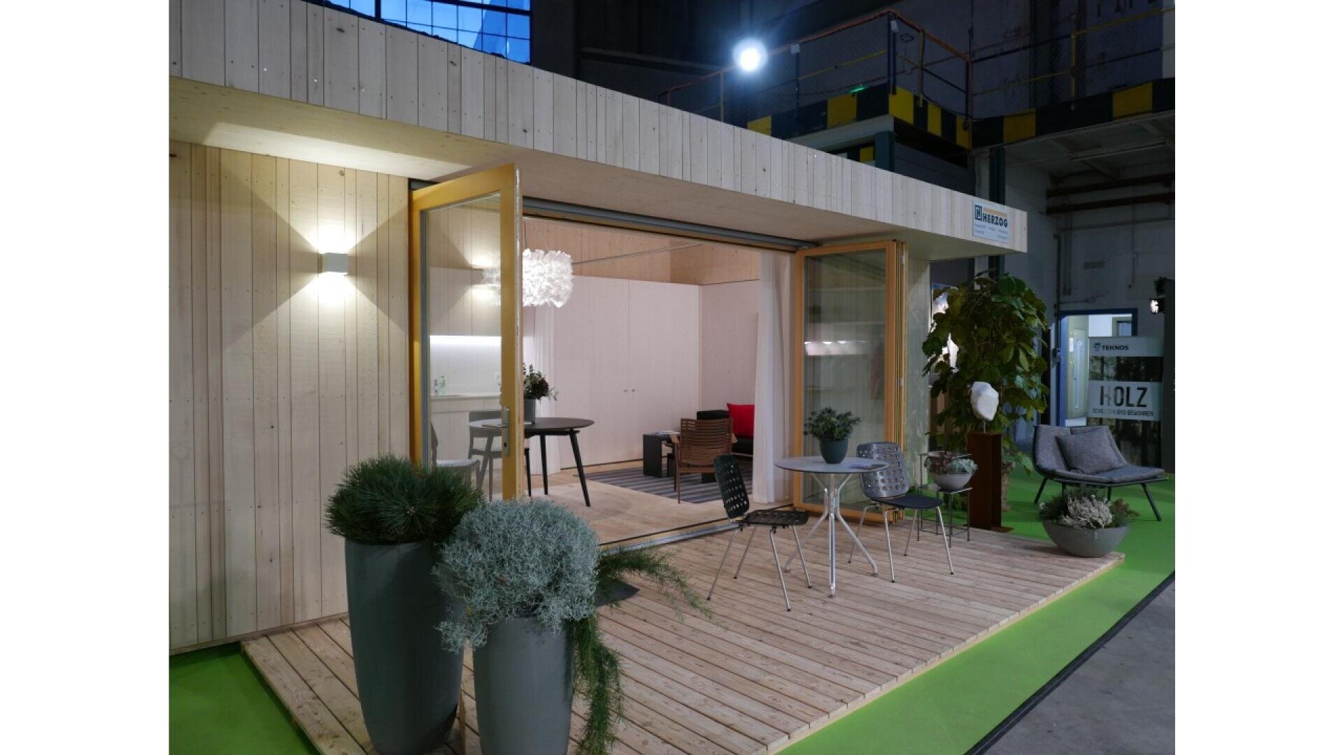 Il s'agit d'une petite maison moderne en bois dont les grandes portes vitrées s'ouvrent sur une petite terrasse agrémentée de plantes en pot, d'une table et de chaises. L'intérieur est très éclairé et meublé simplement. Il semble s'agir d'une exposition en intérieur.