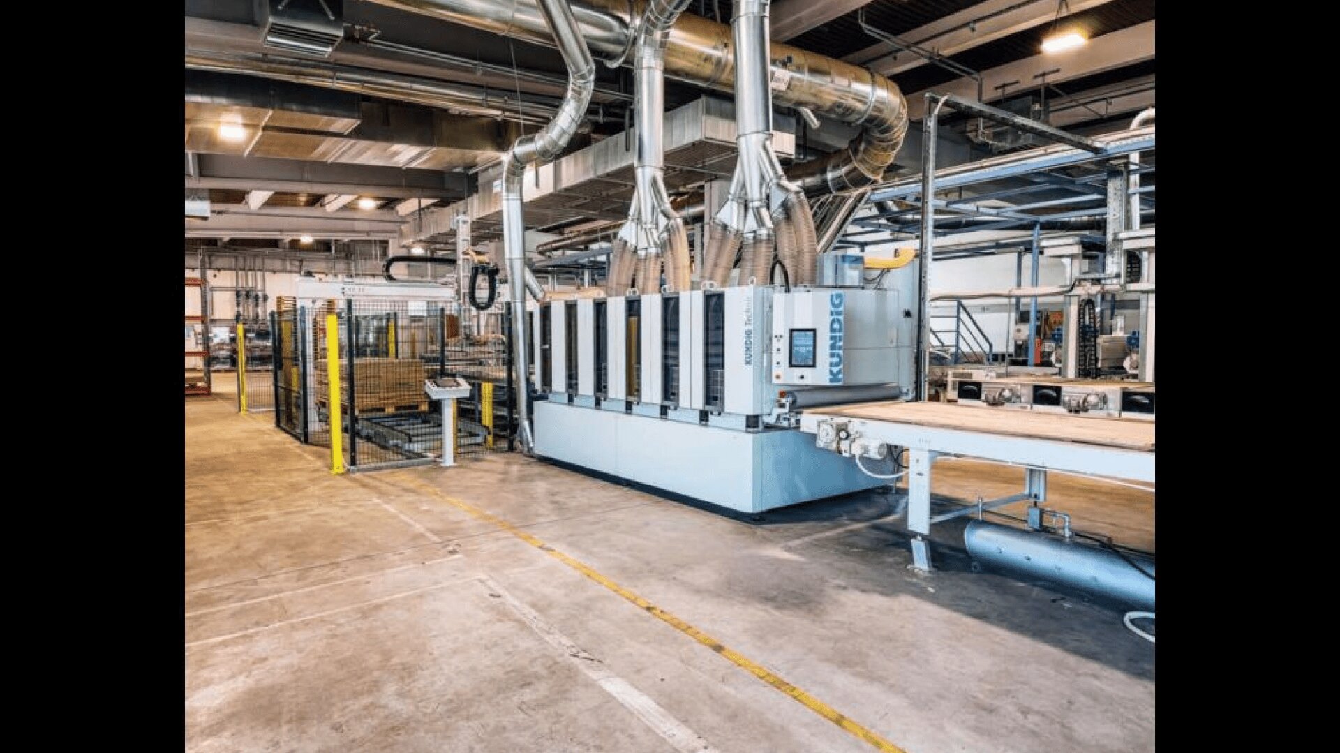 Ein modernes industrielles Fabrikinterieur mit großen automatisierten Maschinen, metallenen Lüftungsrohren und Förderbändern. Der Bereich ist sauber, geräumig und organisiert, mit Metallregalen und verschiedenen Geräten im Hintergrund.