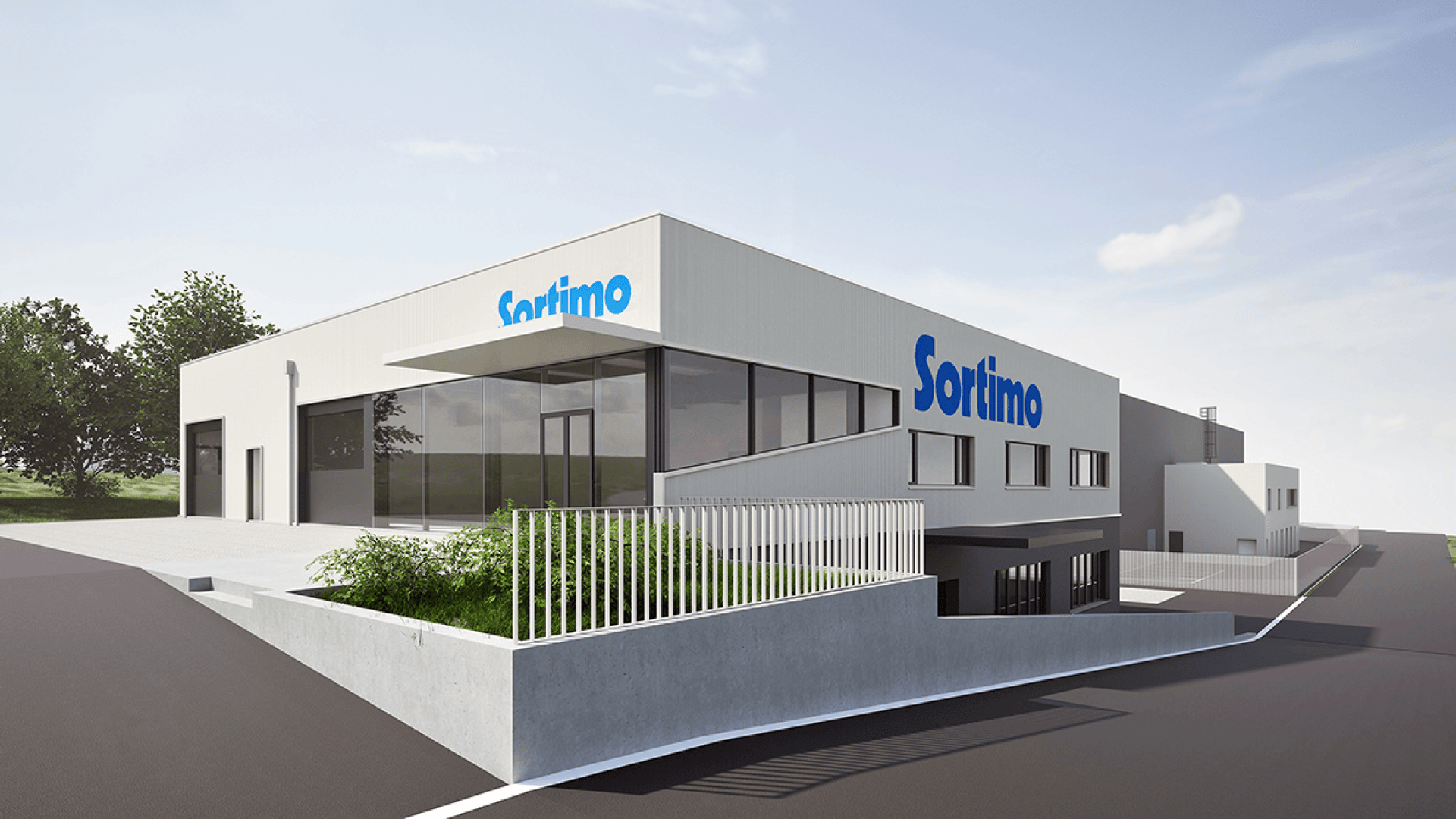 Un bâtiment industriel moderne avec l'inscription Sortimo en bleu sur la façade, avec de grandes fenêtres, une rampe et entouré d'espaces verts et de trottoirs sous un ciel partiellement nuageux.