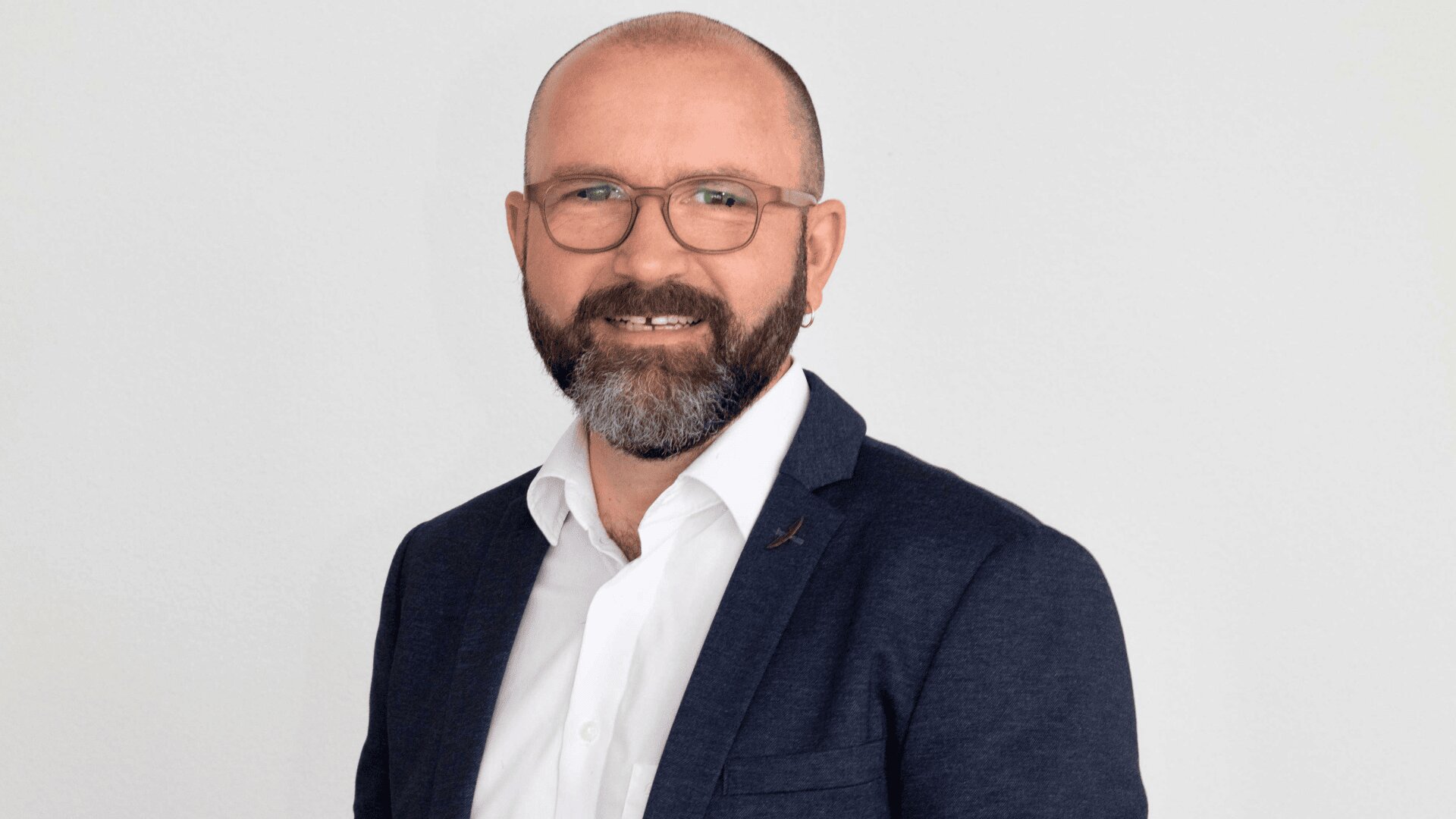 Un homme barbu et portant des lunettes, vêtu d'une chemise blanche et d'un blazer sombre, se tient devant un fond uni de couleur claire et sourit à l'appareil photo.