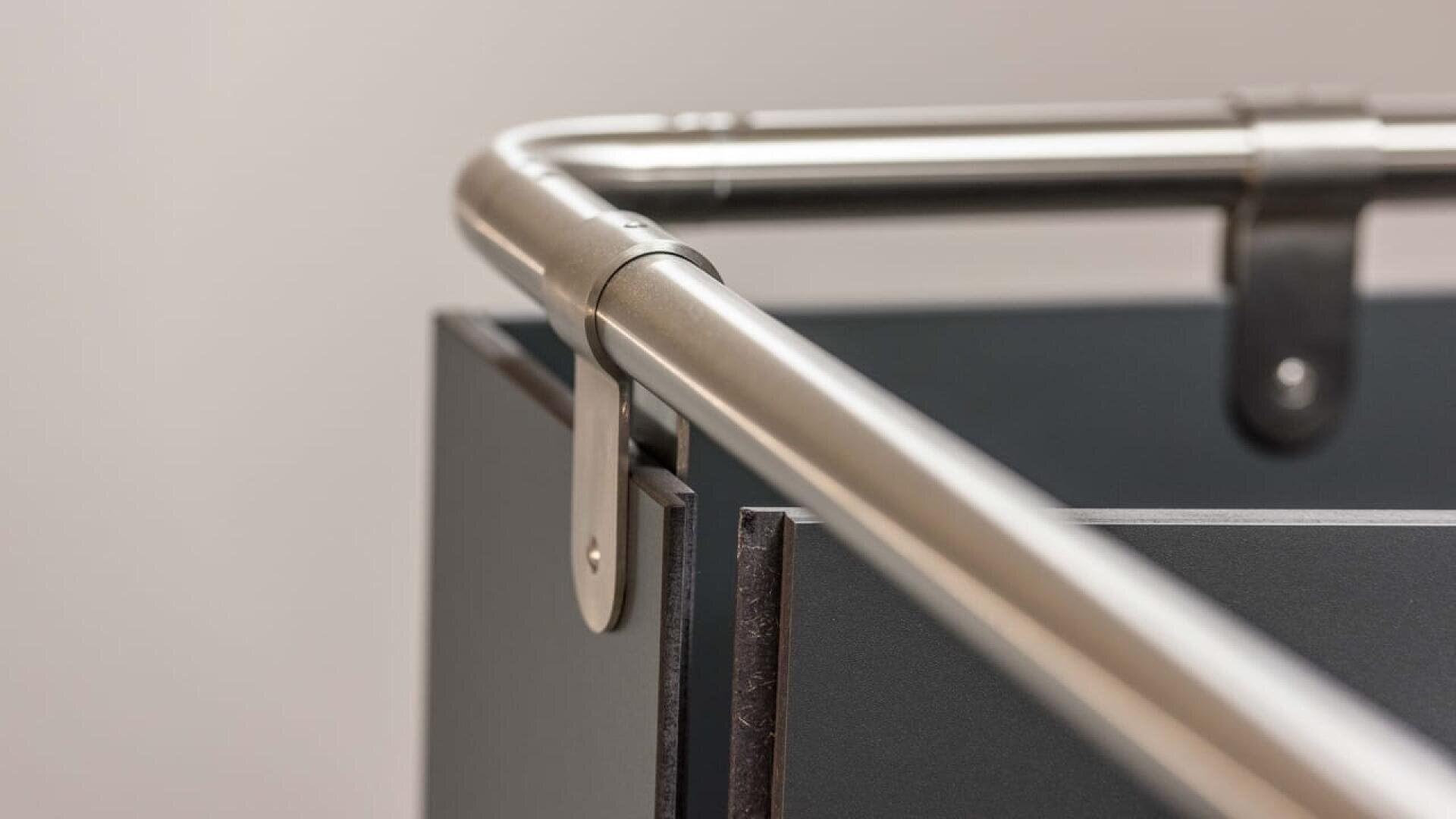 Gros plan d'un rail métallique fixé au sommet d'une structure rectangulaire de couleur foncée, peut-être un meuble ou un conteneur industriel, sur un fond clair uni.