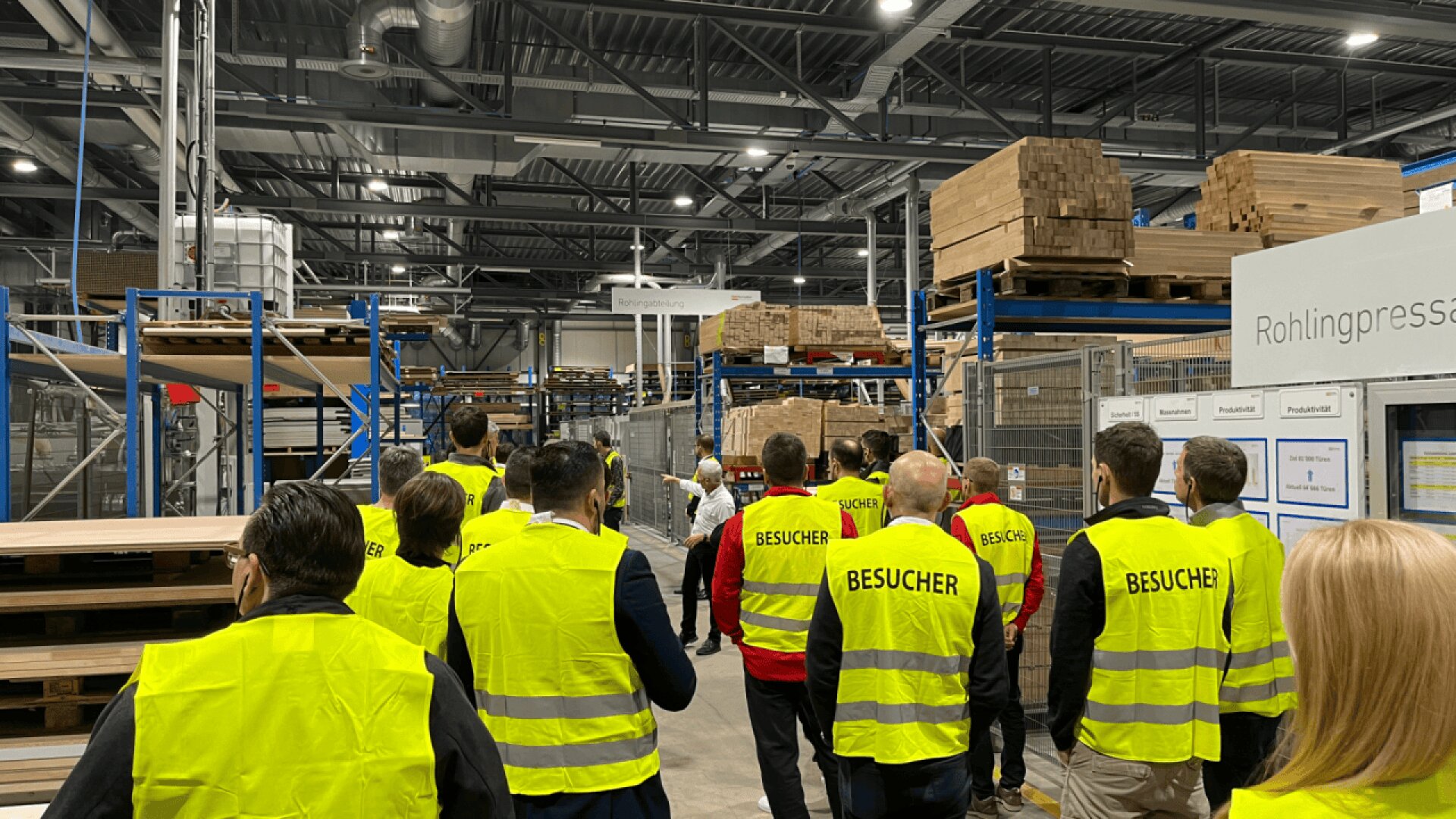 Un groupe de personnes portant des gilets de sécurité jaunes marqués BESUCHER (visiteur) traverse un entrepôt industriel avec des étagères de bois empilées et des machines visibles à l'arrière-plan.