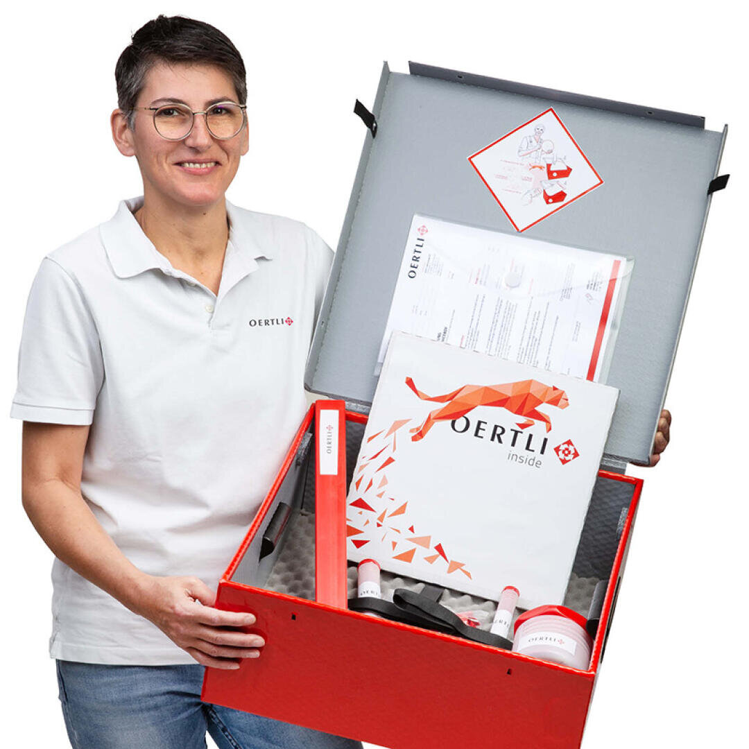 Eine Person mit weissem Poloshirt und Brille lächelt, während sie eine geöffnete rote Schachtel mit Dokumenten, Schläuchen und anderen medizinischen oder technischen Geräten der Marke Oertli in der Hand hält.