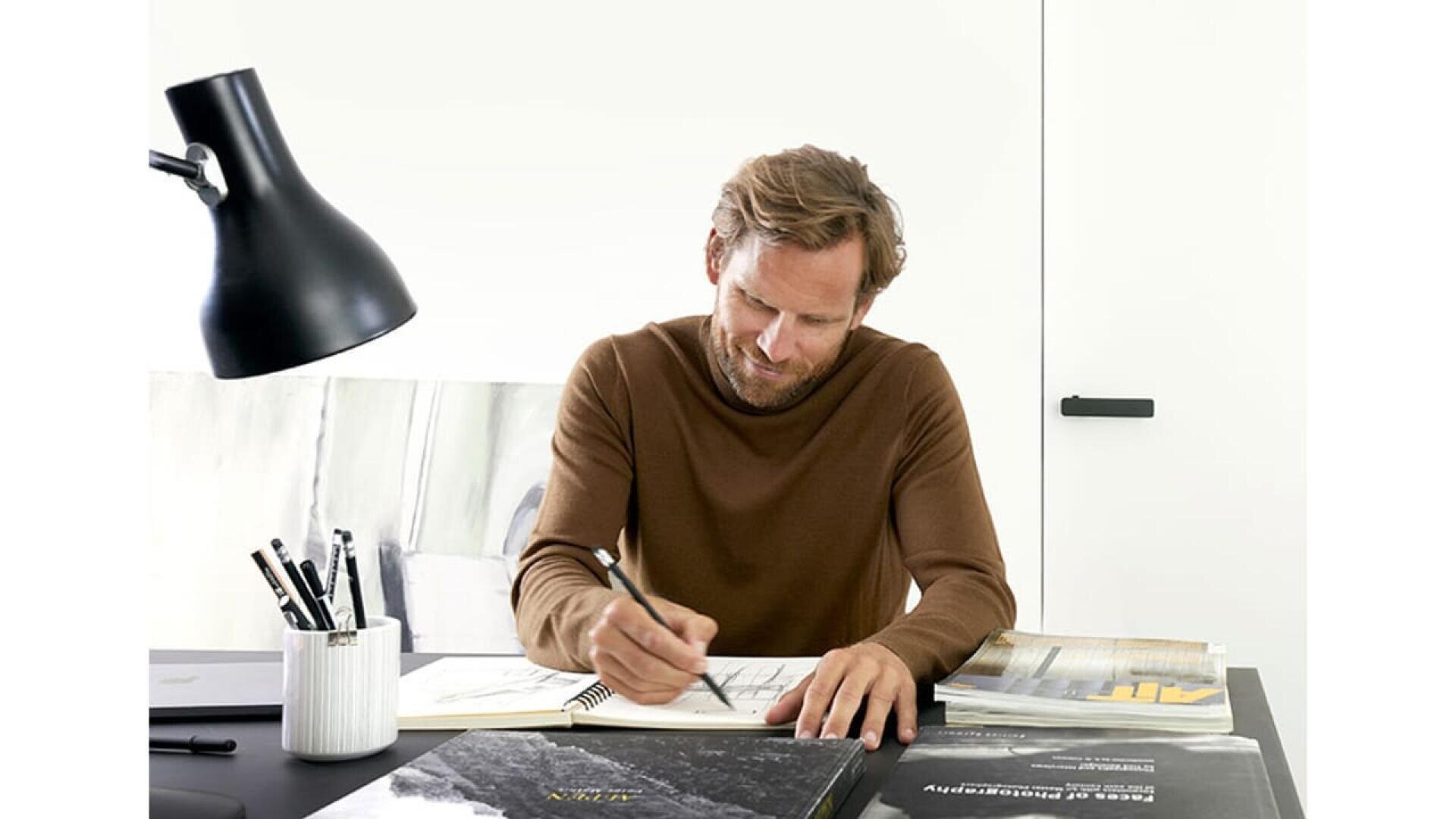 Un homme aux cheveux bruns clairs et à la barbe est assis à un bureau et dessine sur une feuille de papier avec un crayon. Il est entouré de livres, de stylos et d'une lampe de bureau noire dans un espace de travail moderne et lumineux.