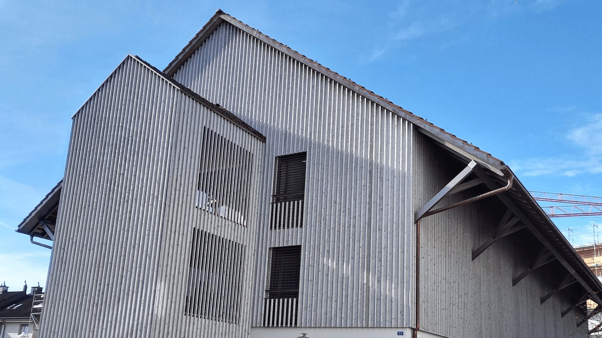 Ein modernes Gebäude mit hellen vertikalen Holzlatten und einem stark geneigten Dach steht vor einem klaren blauen Himmel; einige Fenster sind teilweise von den Latten verdeckt, und im Hintergrund ist ein weiteres Gebäude zu sehen.