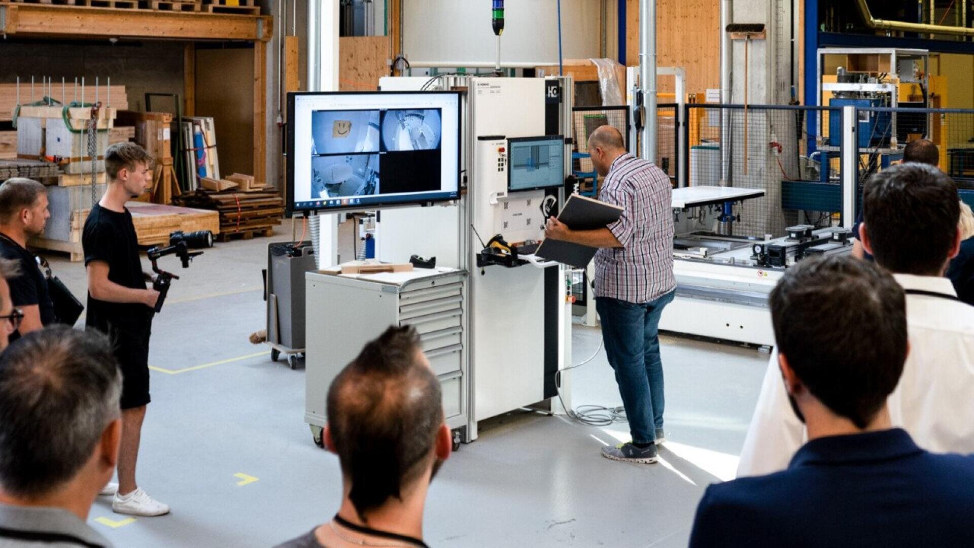 Un groupe de personnes observe un homme qui utilise une machine avec deux écrans dans un vaste atelier industriel. Certains écrans affichent des images techniques, tandis que des équipements et des matériaux sont visibles à l'arrière-plan.